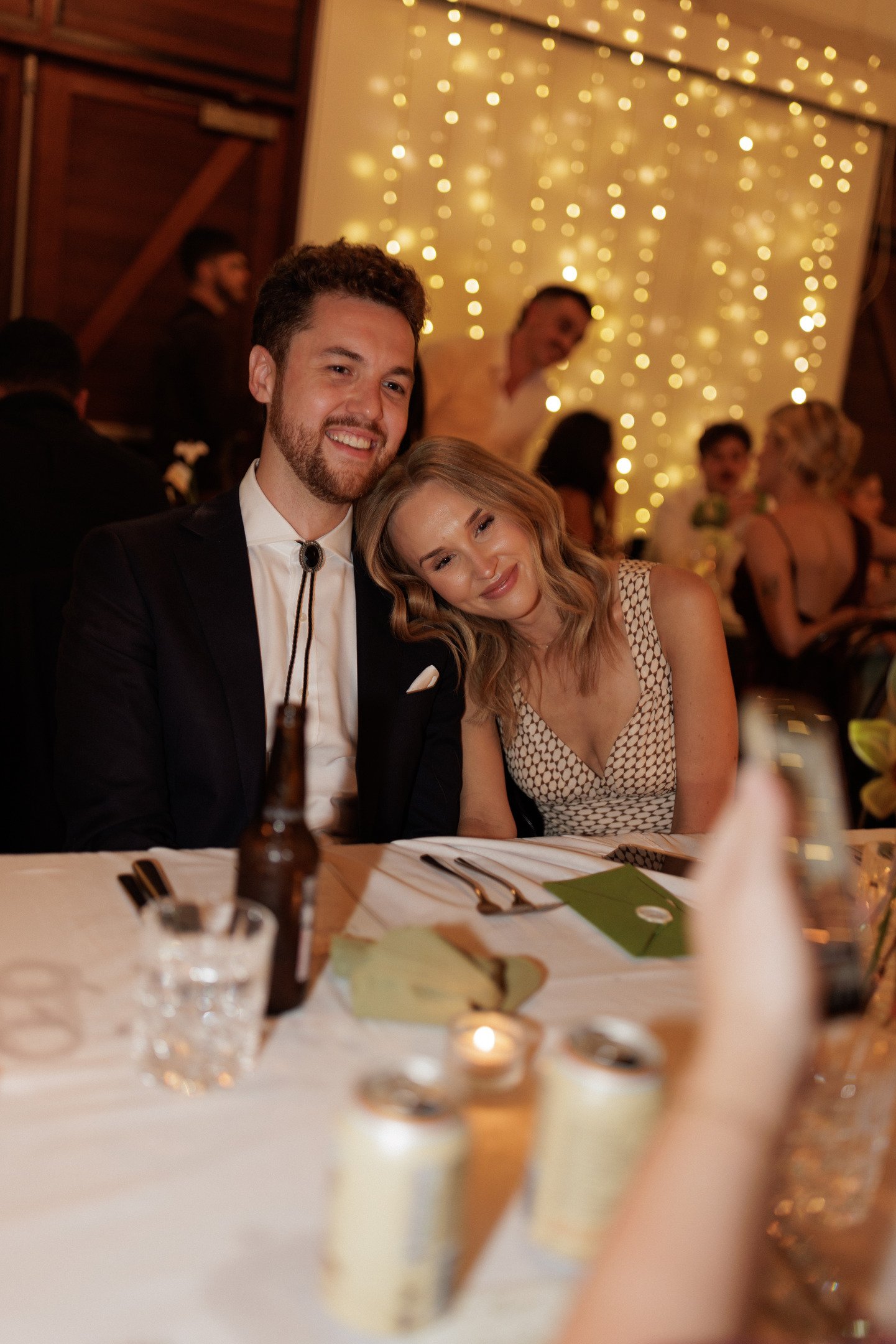A man in a tuxedo and a woman in a patterned dress sitting at a decorated table during a celebration, with blurred background of people and string lights.