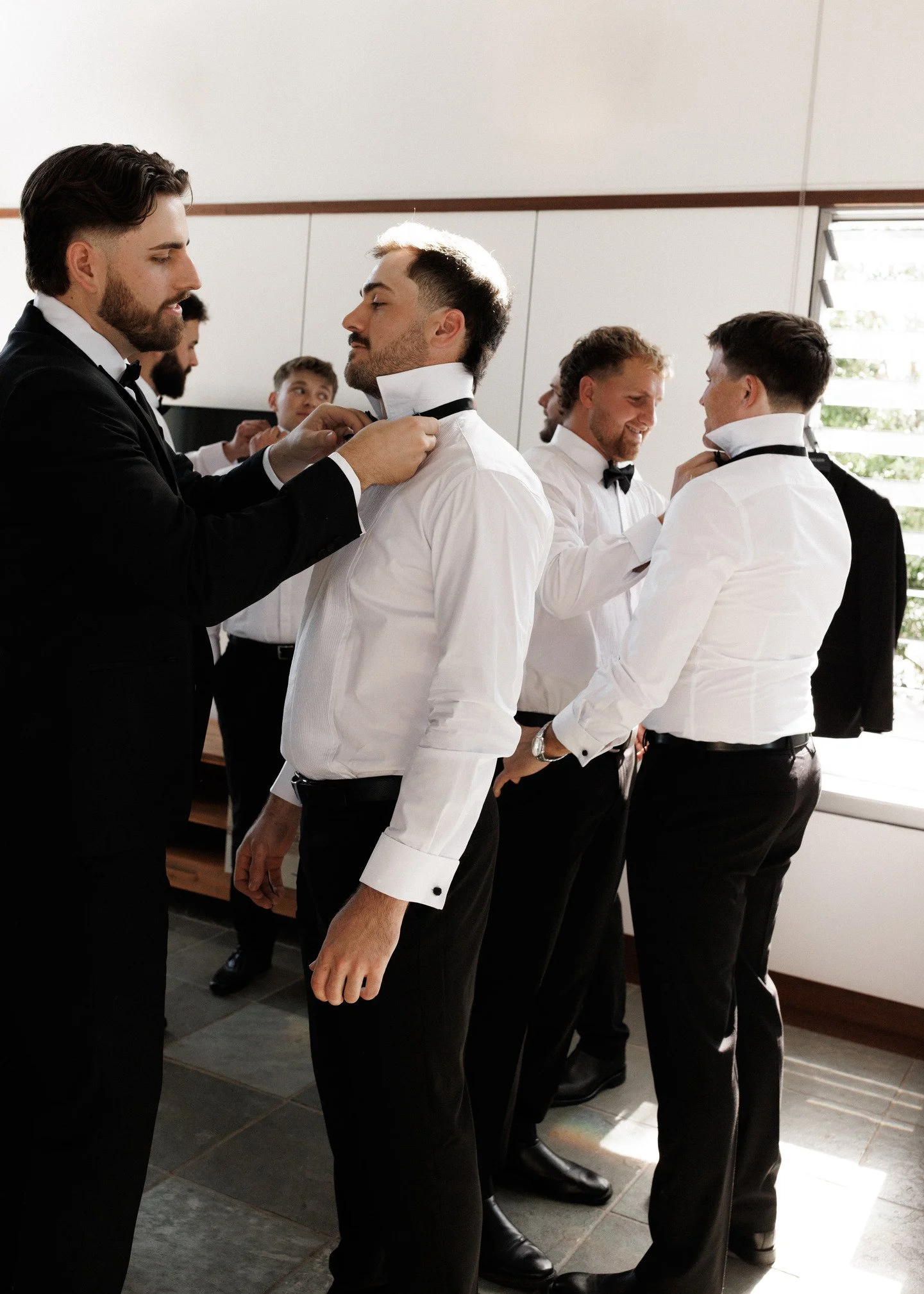 Group of men dressed in tuxedos and white shirts, standing in a line, adjusting their bow ties and collars, preparing for a formal event in a well-lit room with large windows.