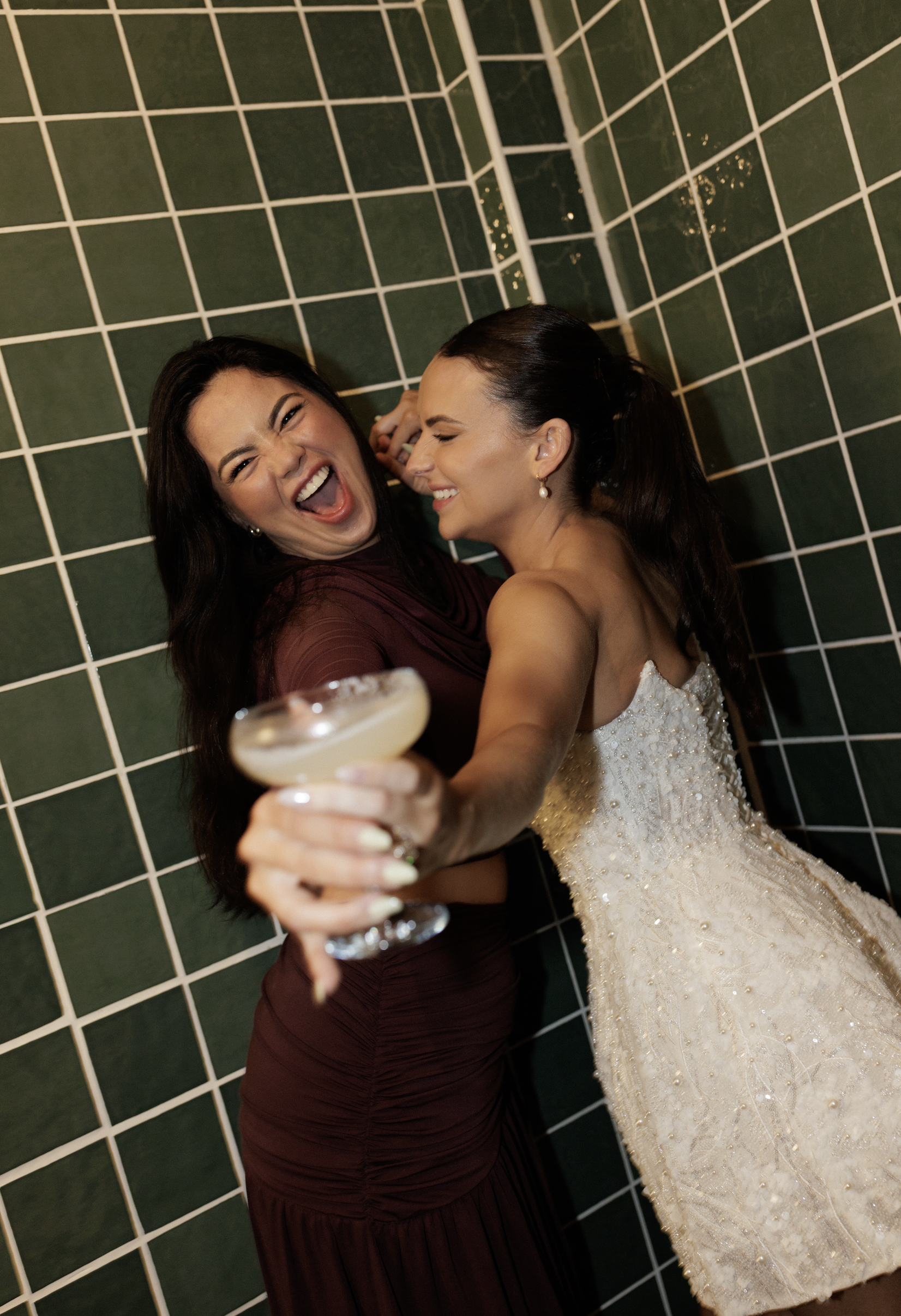 Two women smiling and taking a selfie in front of a green tiled wall, one dressed in a wedding gown and the other in a dark dress, holding a cocktail glass.