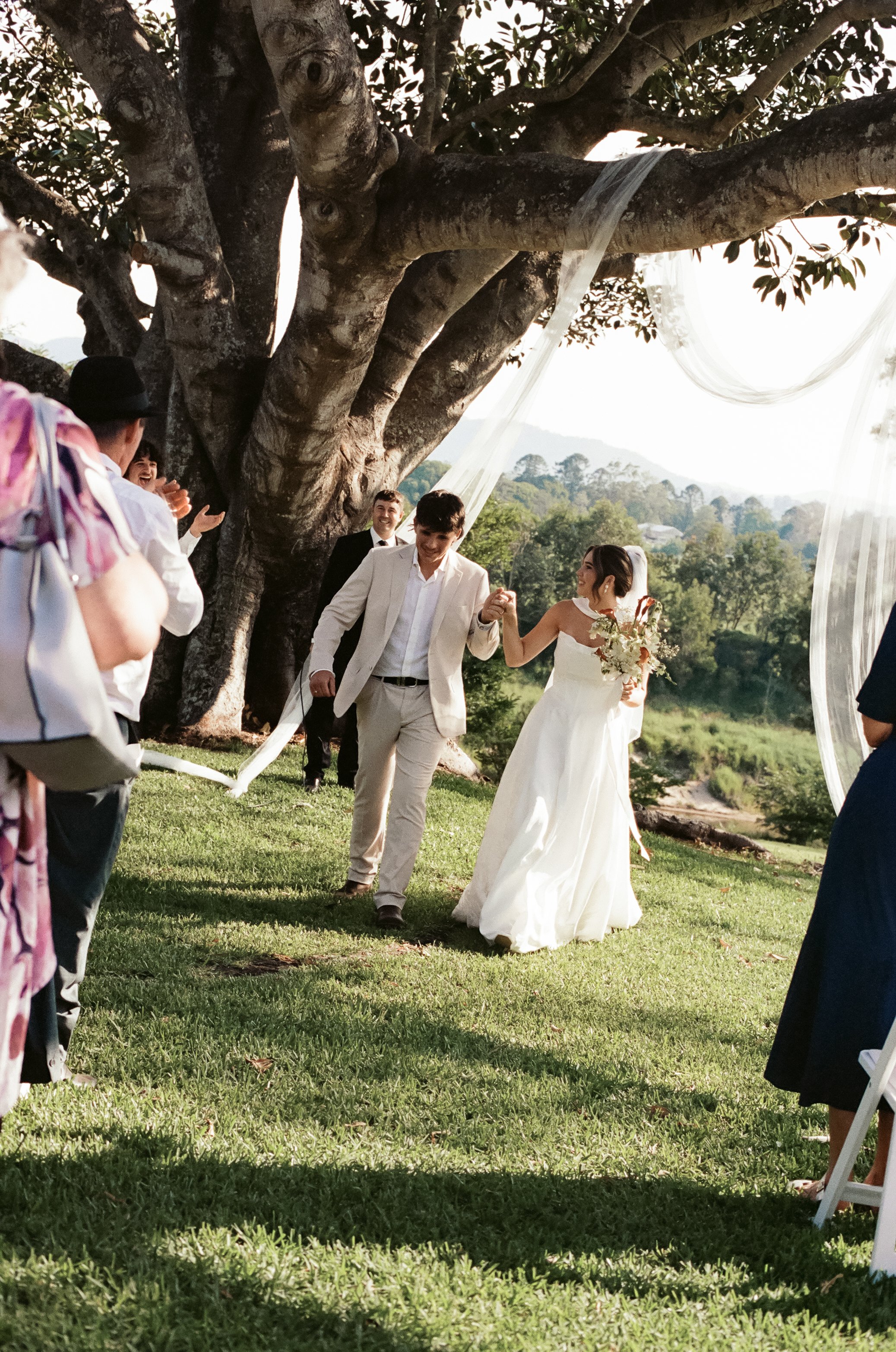 A bride and groom walking hand in hand under a large tree during their outdoor wedding ceremony with guests clapping nearby.
