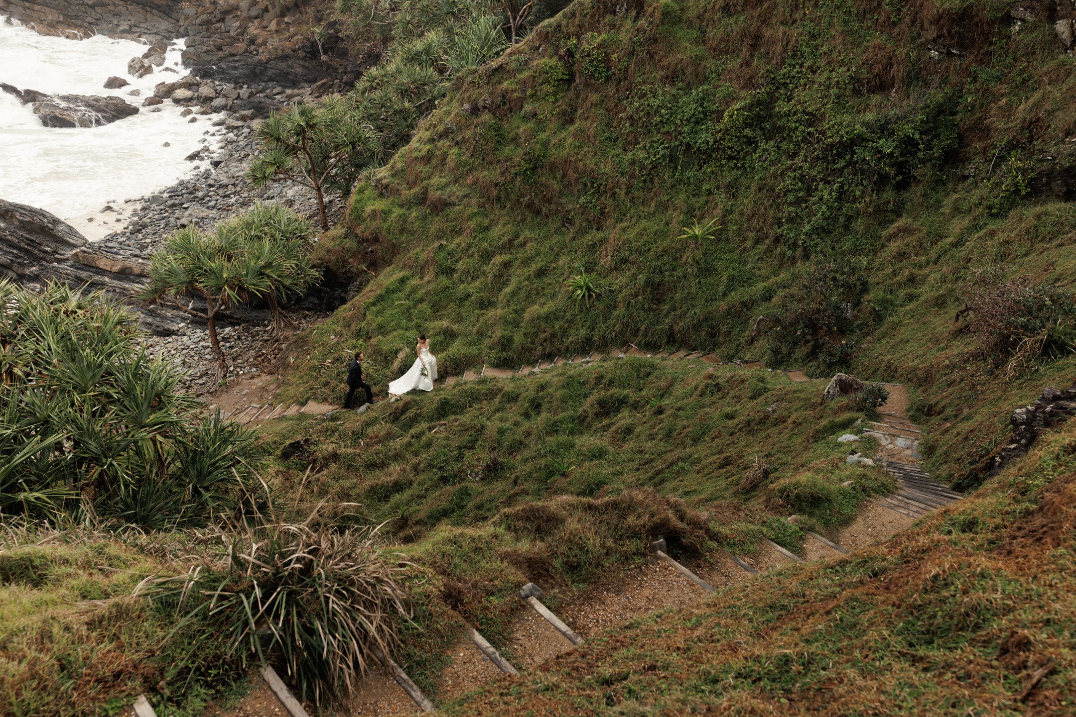 A wedding couple on a winding hillside trail with lush greenery and trees, near a rocky coastline with waves in the background.