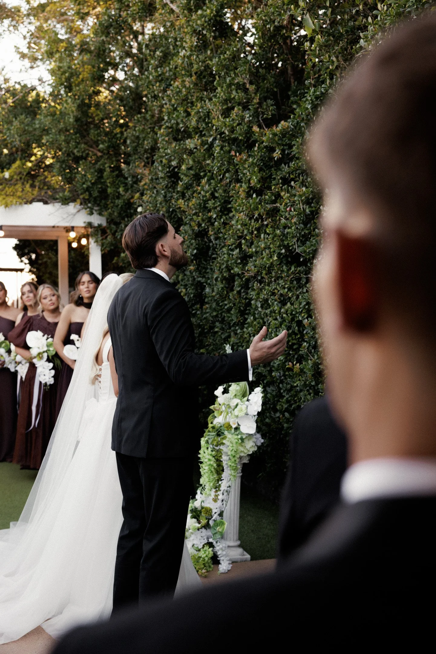 A wedding ceremony with a man in a black suit and a woman in a white wedding dress standing before a green leafy wall, with bridesmaids in burgundy dresses in the background, outdoors during daytime.