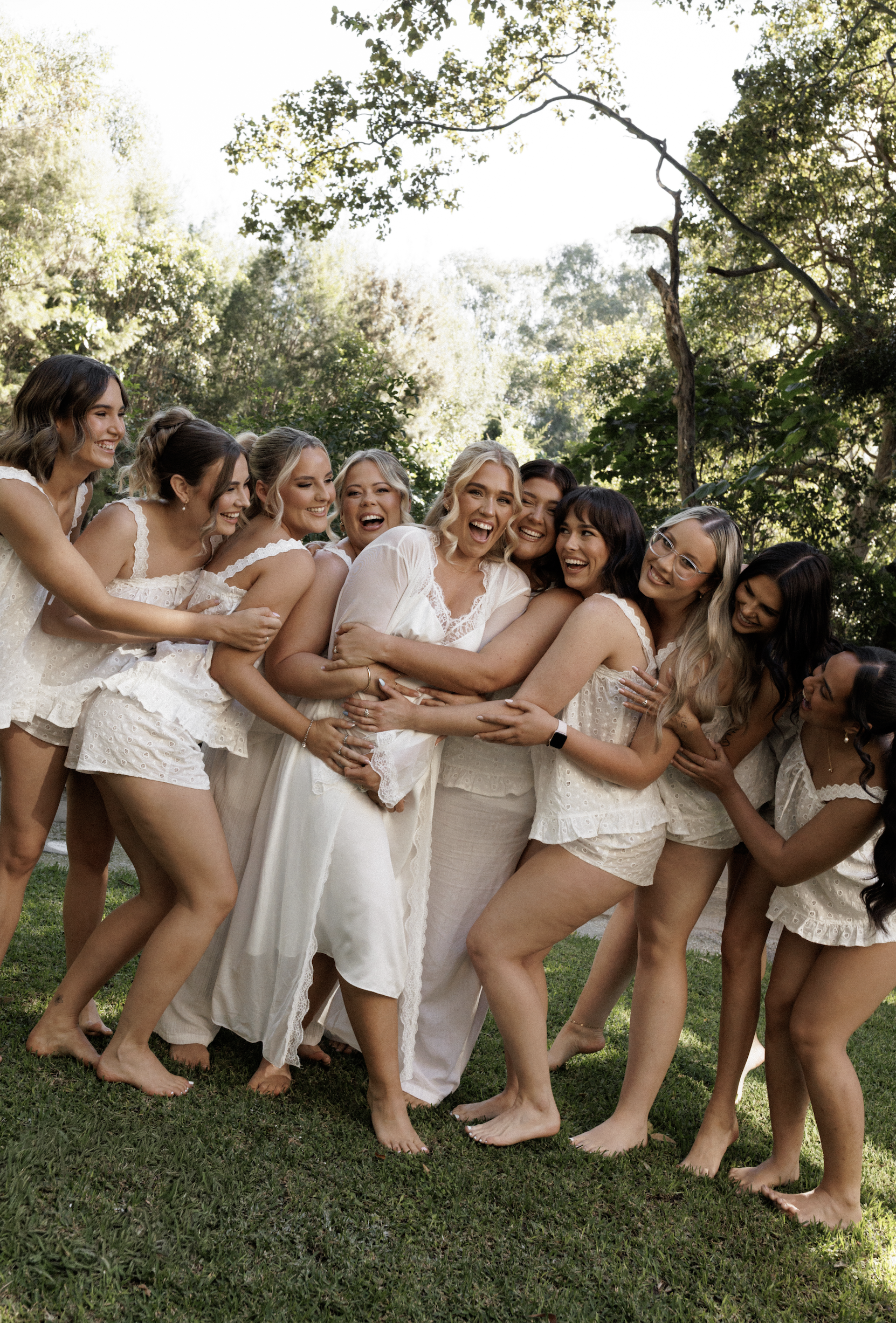 A group of women in white dresses hugging and laughing outdoors on grass with trees and sunlight in the background.