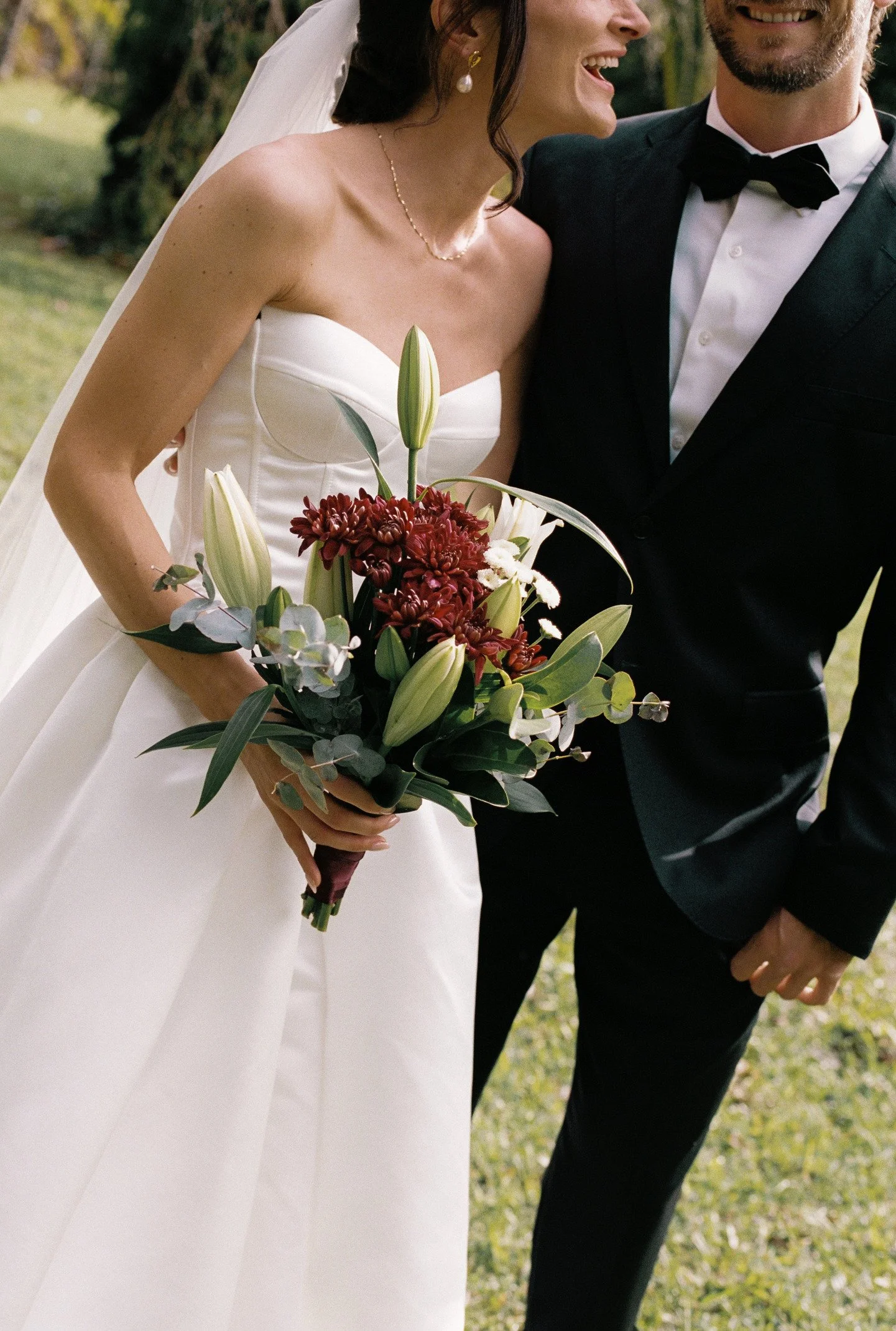 Bride in a white wedding dress holding a bouquet of red and white flowers, smiling, standing next to groom in a black tuxedo with bow tie, outside on grass.