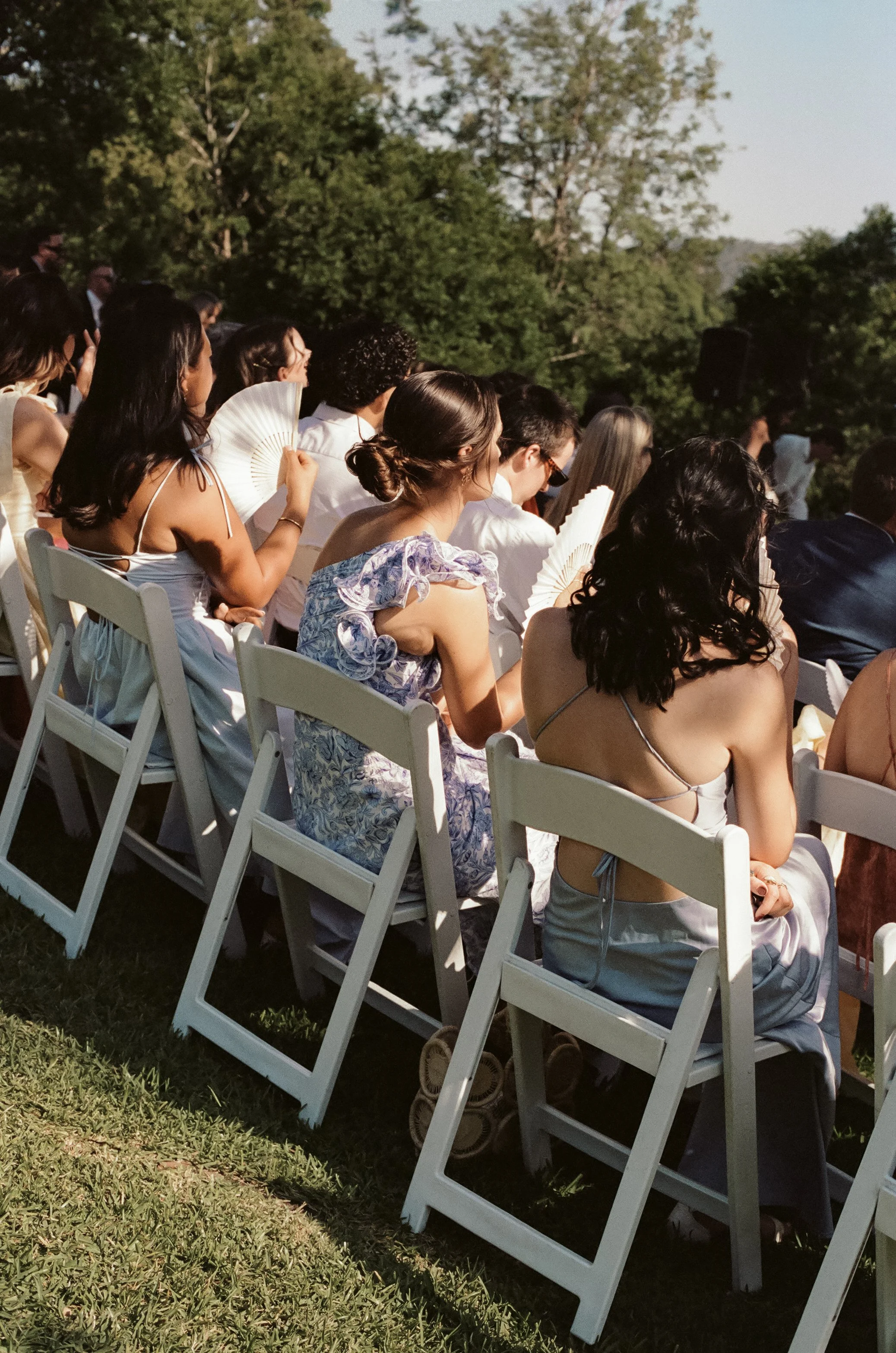 People attending an outdoor event, sitting on white chairs with some holding fans, on a grassy area surrounded by trees.