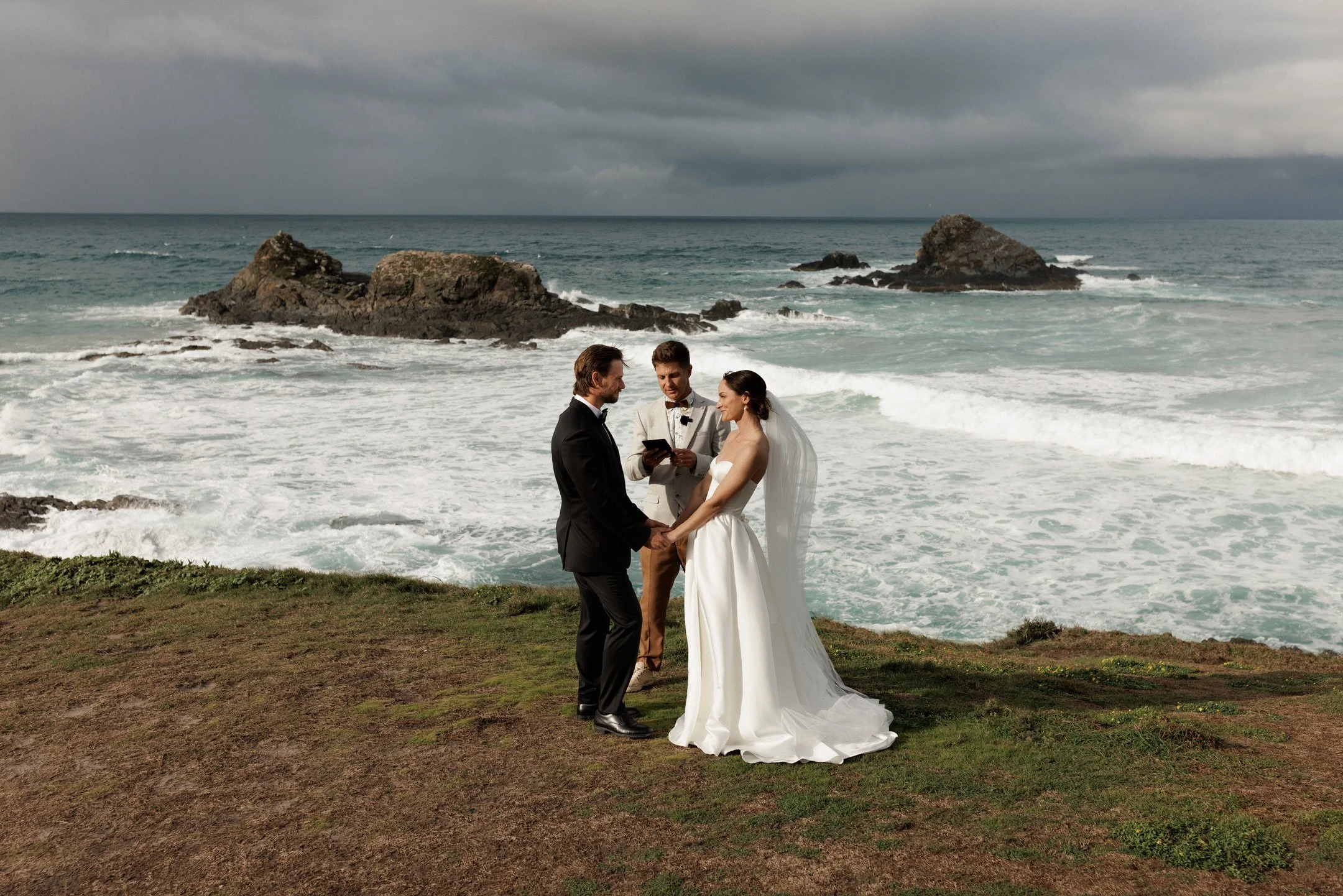 A couple in wedding attire exchanging vows on a grassy area near the ocean, with a rocky shoreline and cloudy sky in the background.