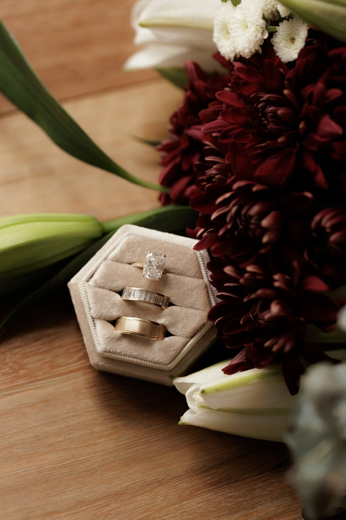 Close-up of a jewelry box with four rings, placed among white and dark red flowers on a wooden surface.