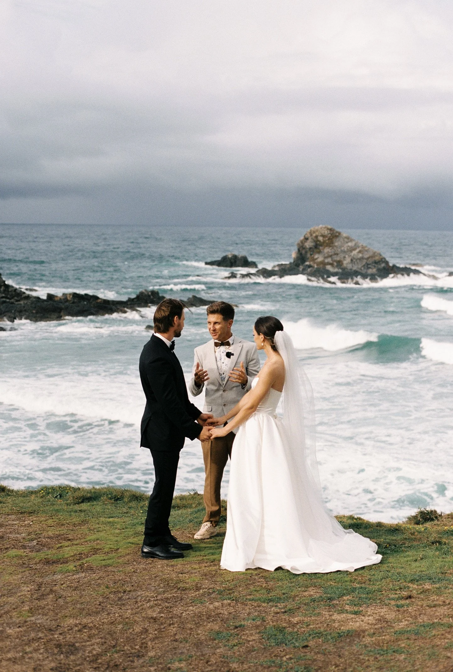 A couple getting married on a grassy area near the beach, with an officiant during the ceremony. The couple is holding hands and facing each other, with a rocky ocean view and cloudy sky in the background.