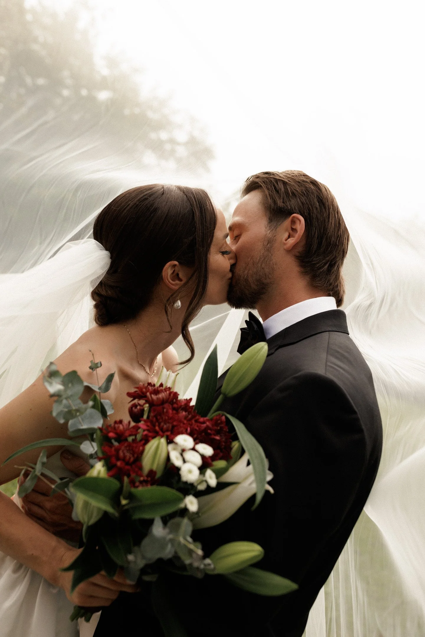 A bride and groom kiss at their wedding, the bride holding a bouquet of red, white, and green flowers, under a sheer fabric canopy outdoors.