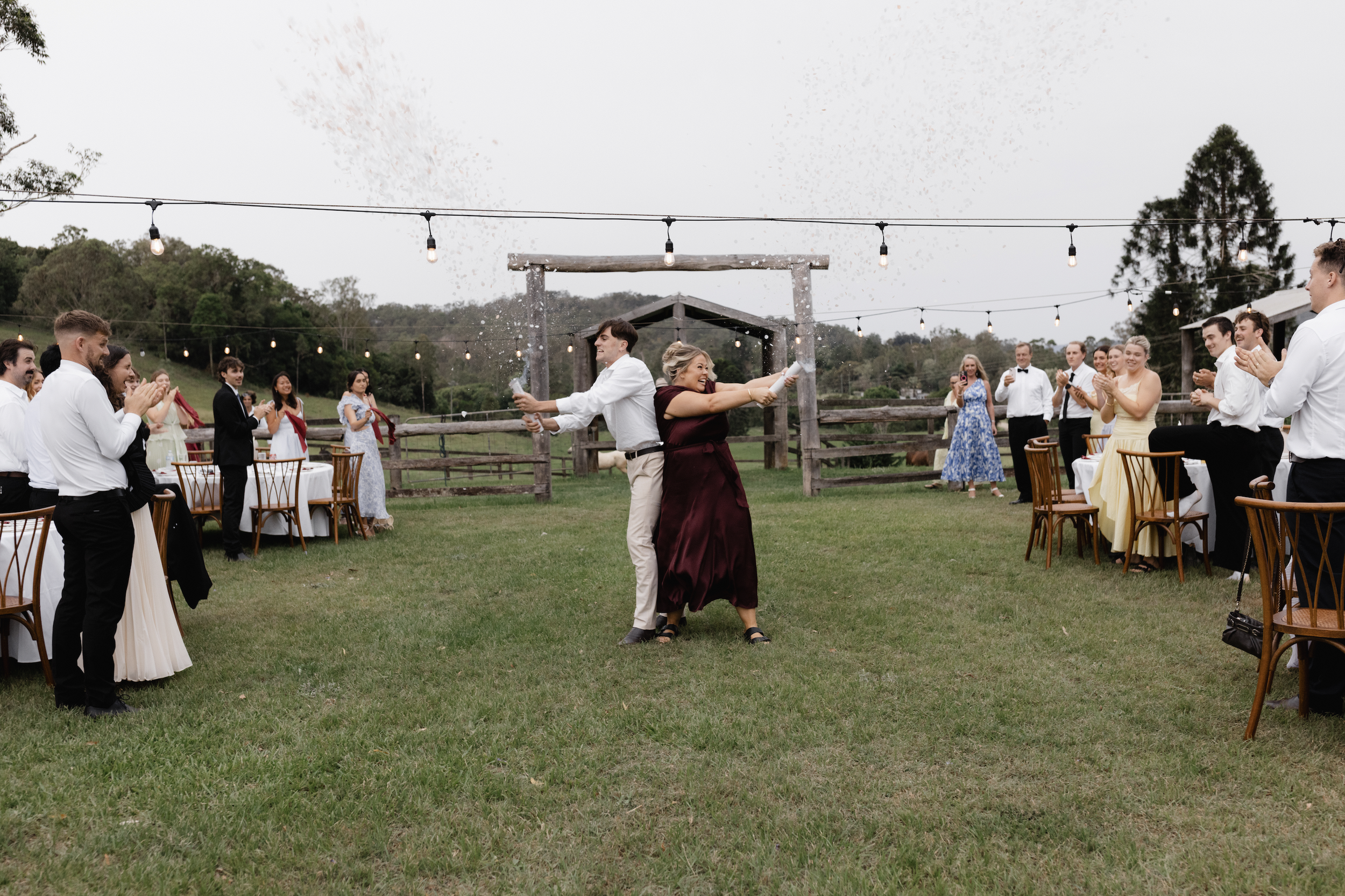 People celebrating at an outdoor wedding reception, with two individuals in the center popping champagne and people around clapping and smiling.