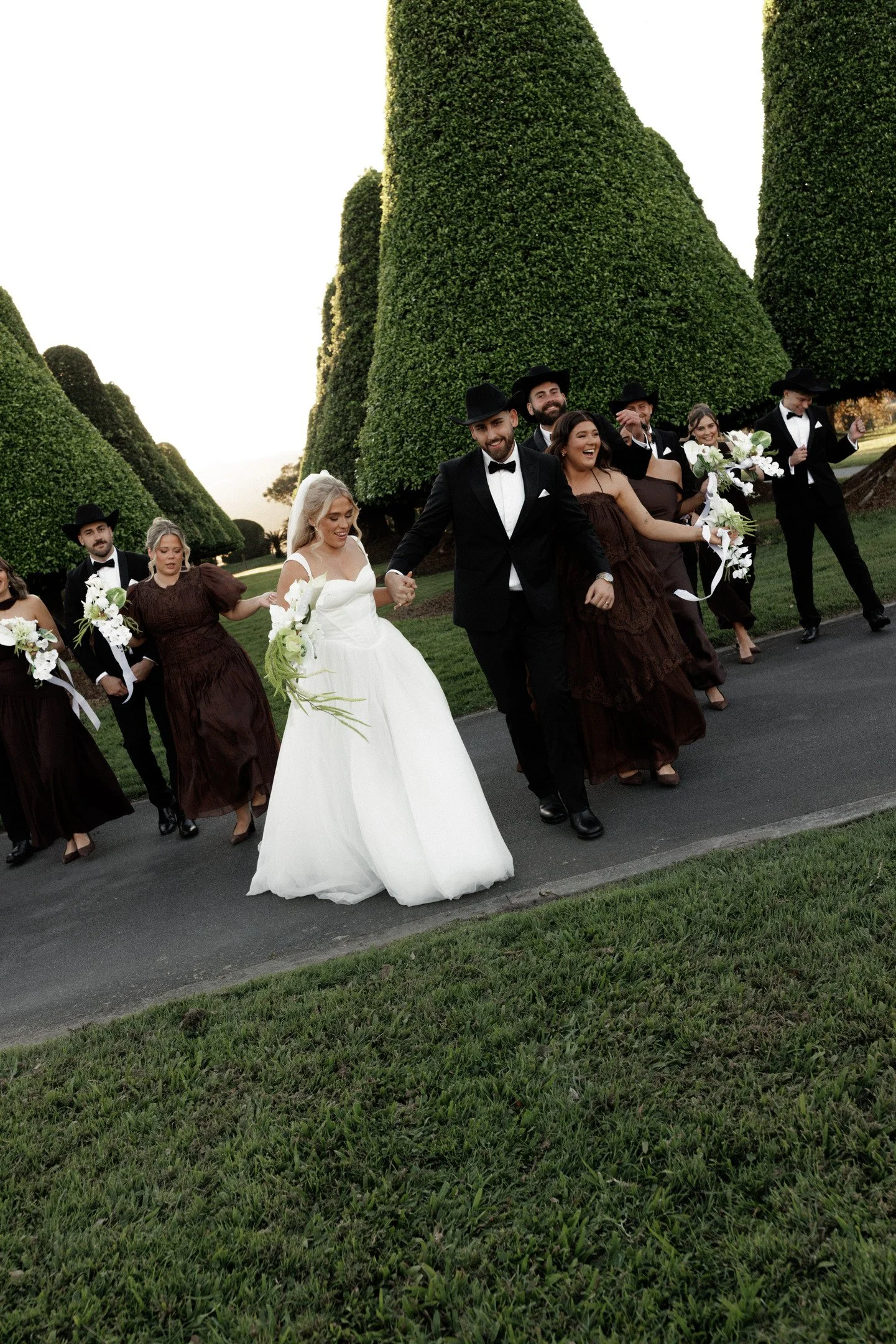 A group of people dressed formally, celebrating and walking outdoors on a paved pathway surrounded by large, well-manicured topiary trees. The women are in brown dresses and the men in tuxedos with black hats, two women are holding bouquets of white 