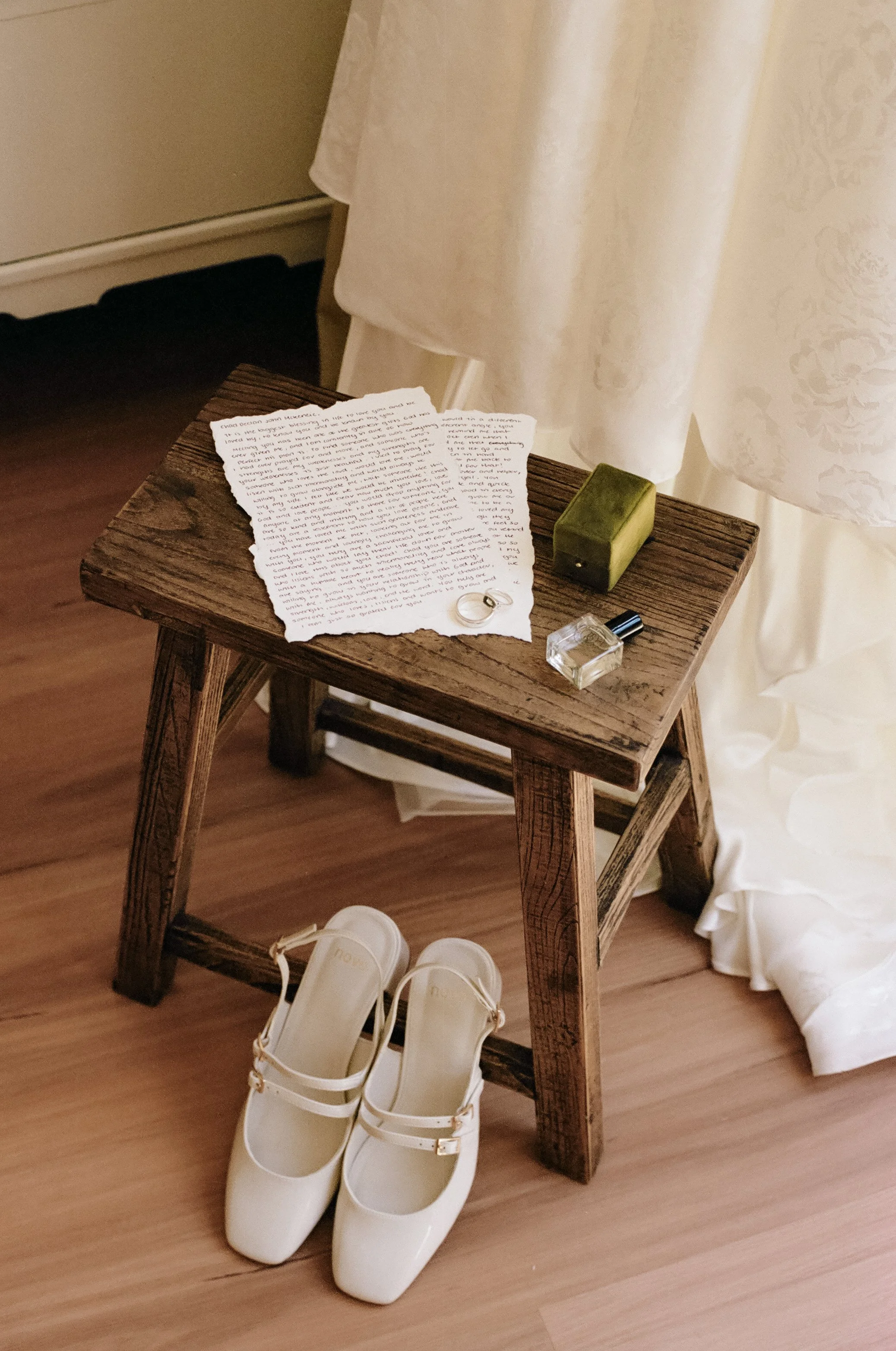 A small wooden table with wedding accessories: a wedding ring, a green box, a perfume bottle, and a nail polish bottle. A pair of white dress shoes is placed underneath the table. The table is near cream-colored curtains.