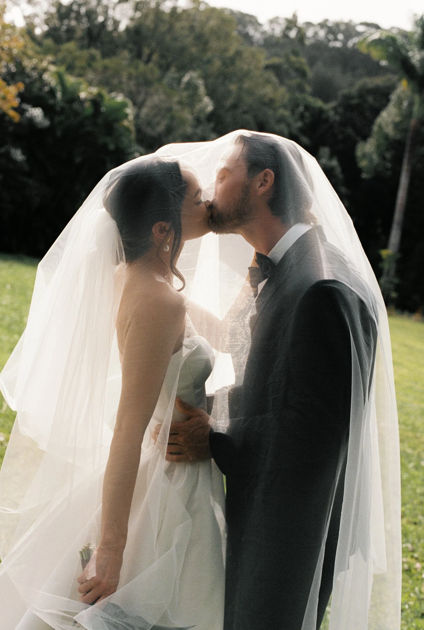 A bride and groom share a kiss beneath a sheer wedding veil outdoors on a sunny day, with trees and greenery in the background.