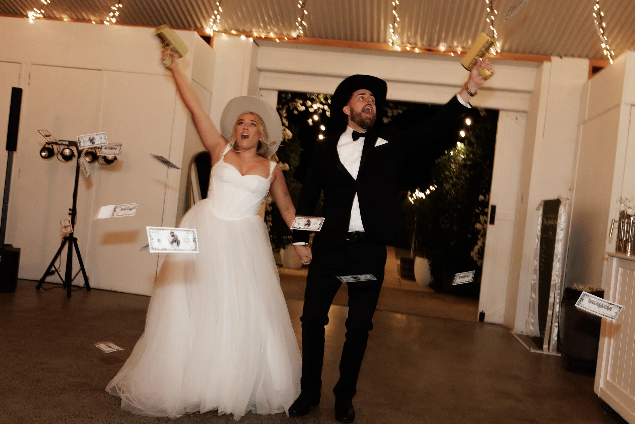 A bride and groom celebrating at their wedding reception, holding hands and raising glasses of champagne. The bride is wearing a white wedding gown, and the groom is dressed in a black tuxedo with a black hat. There are string lights hanging from the