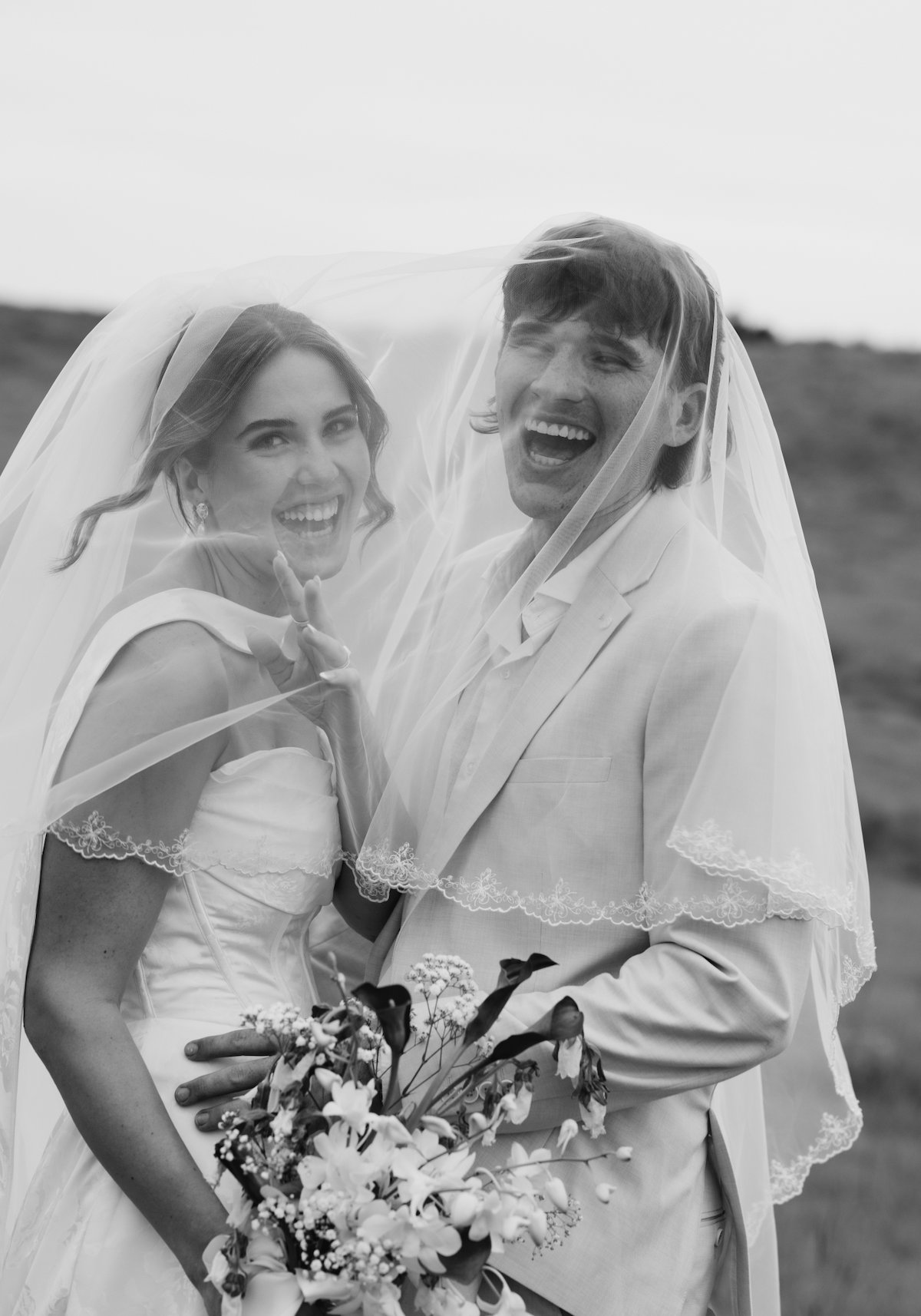 A happy bride and groom under a veil, smiling outdoors with a bouquet of flowers.