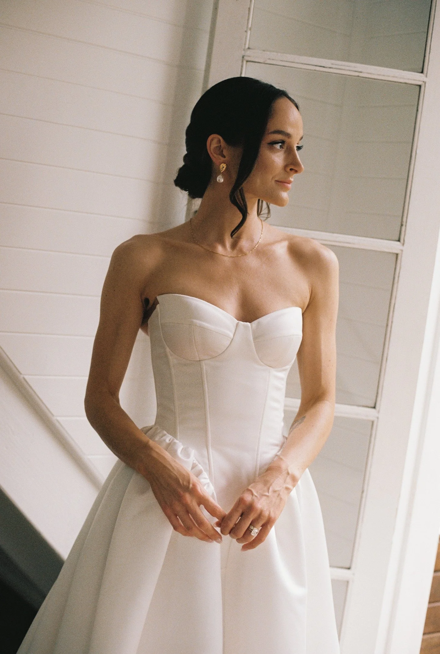A woman in a strapless white wedding dress is looking to the right, standing indoors near a mirror with white panels on the wall behind her.