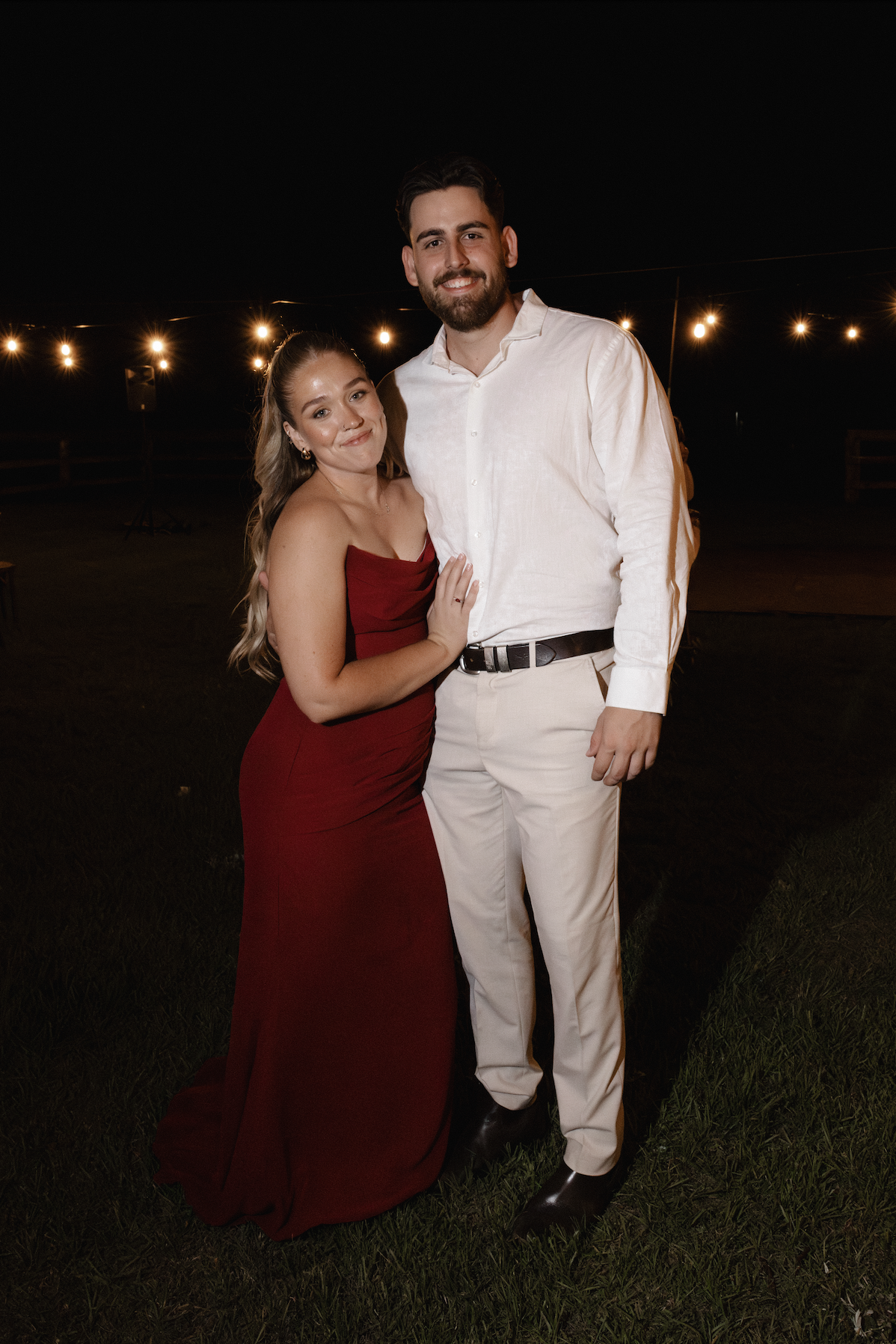 A young couple dressed in formal attire standing outdoors at night with string lights in the background. The woman is wearing a red strapless dress and the man is wearing a white shirt and light-colored pants.