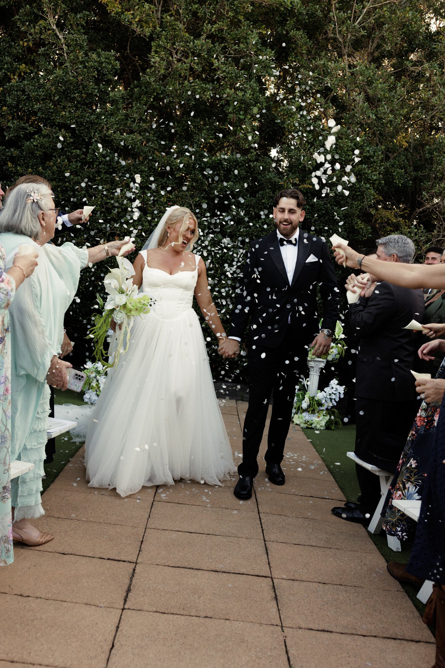 A wedding ceremony outdoors with a bride and groom holding hands while guests throw white flower petals. The bride wears a white wedding gown and veil, holding a bouquet, and the groom wears a black tuxedo with a bow tie. They are standing on a path 