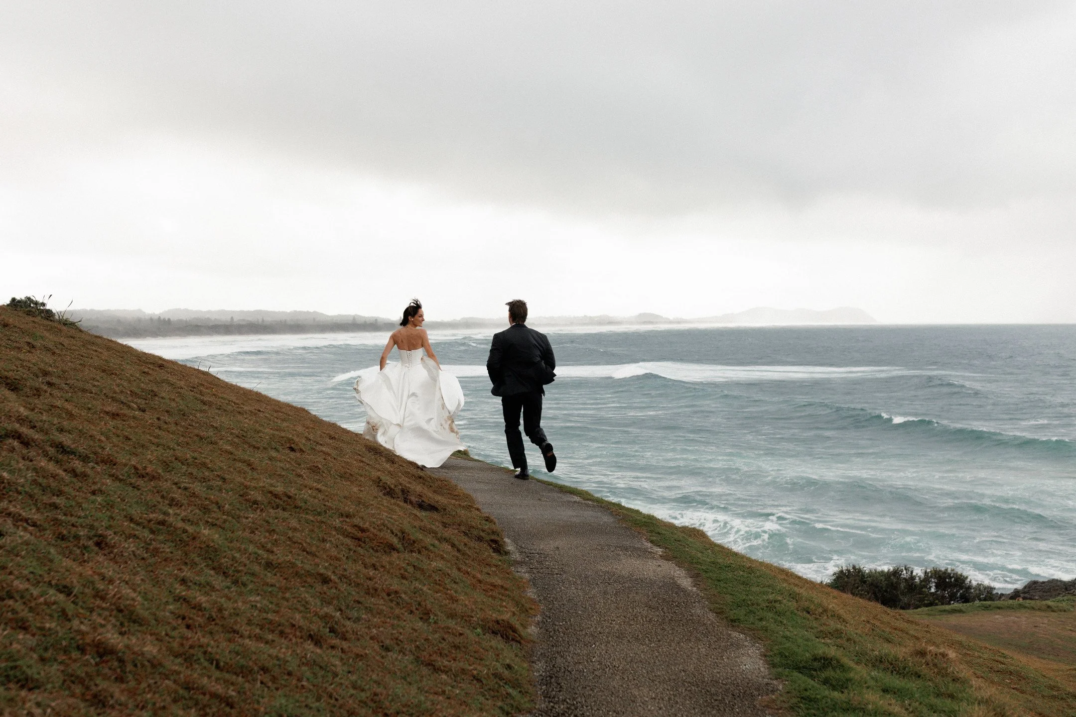 A bride and groom walking along a coastal path near the ocean on a cloudy day.