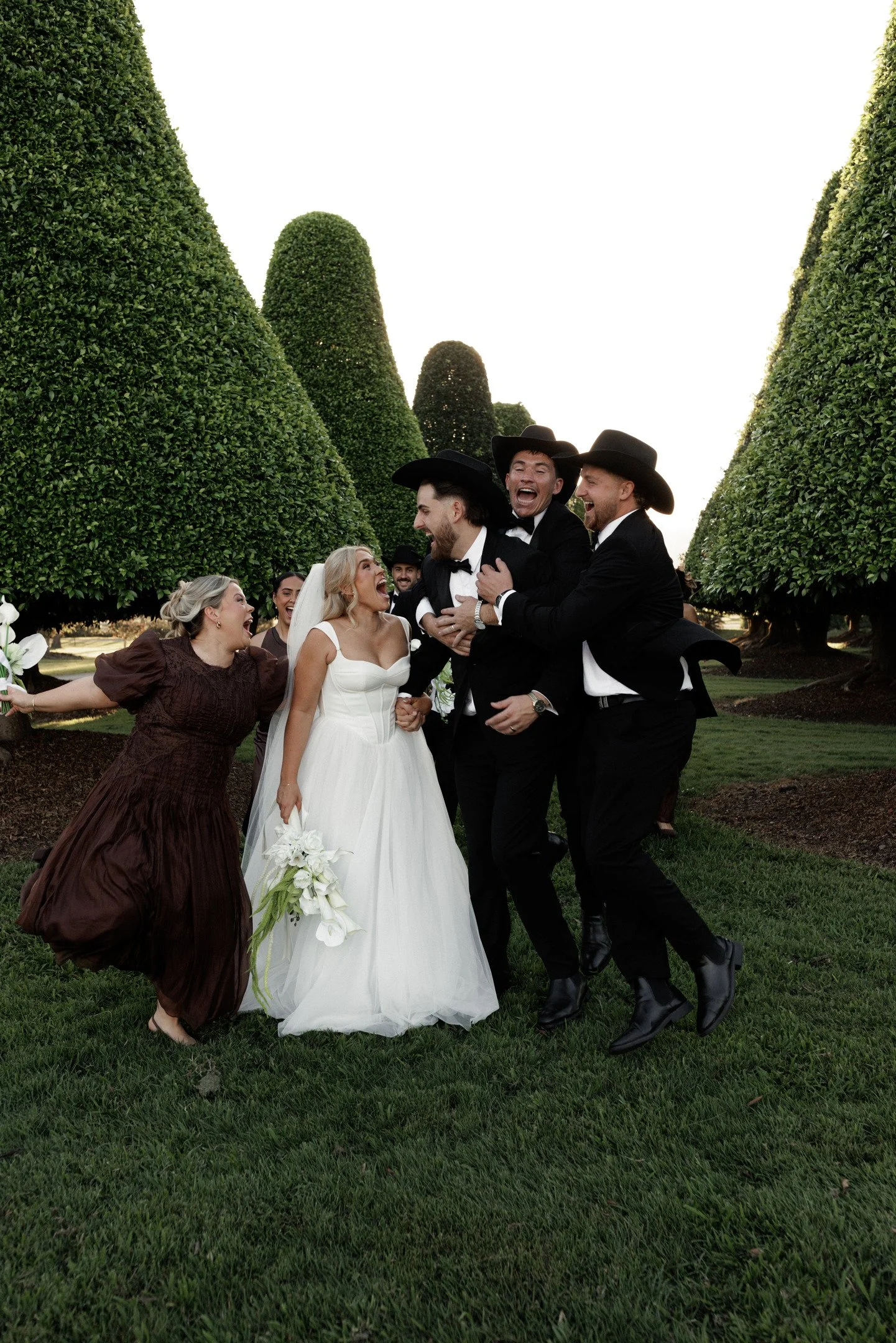 A group of people celebrating a wedding outdoors among tall, manicured trees, including a bride in a white dress and several men in black suits and cowboy hats, all smiling and laughing.