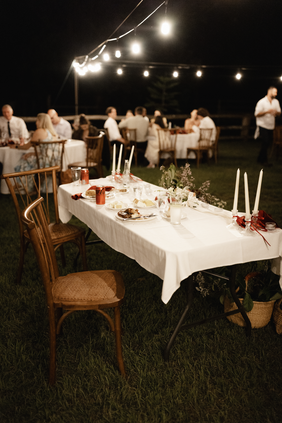 Outdoor evening wedding or party reception with decorated tables, string lights, and guests seated at tables in the background.
