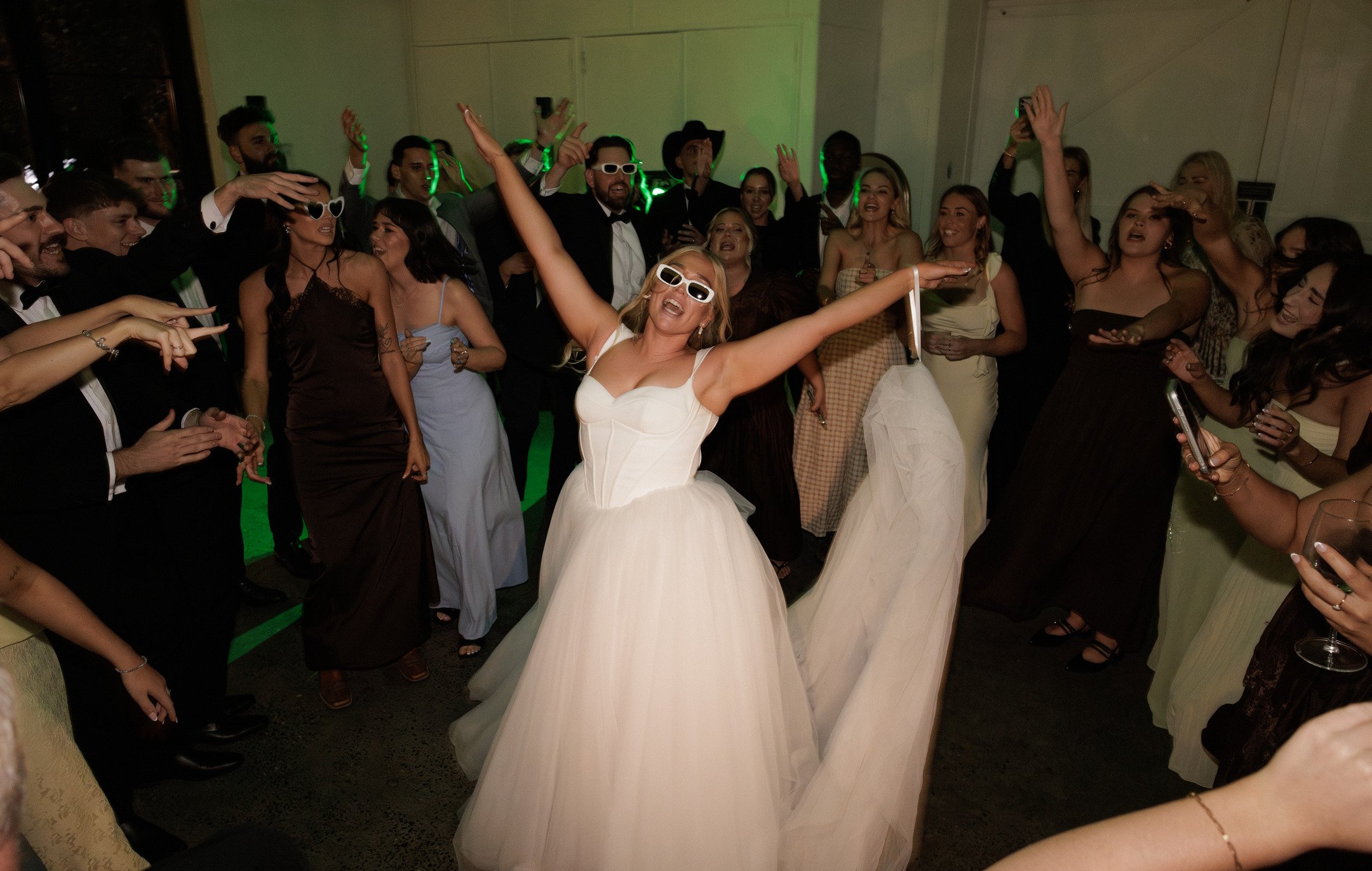 A woman in a white wedding dress and sunglasses dancing with arms raised at a wedding reception. Surrounded by guests in formal attire, some wearing glasses, raising hands, and smiling. The scene is lively and celebratory.