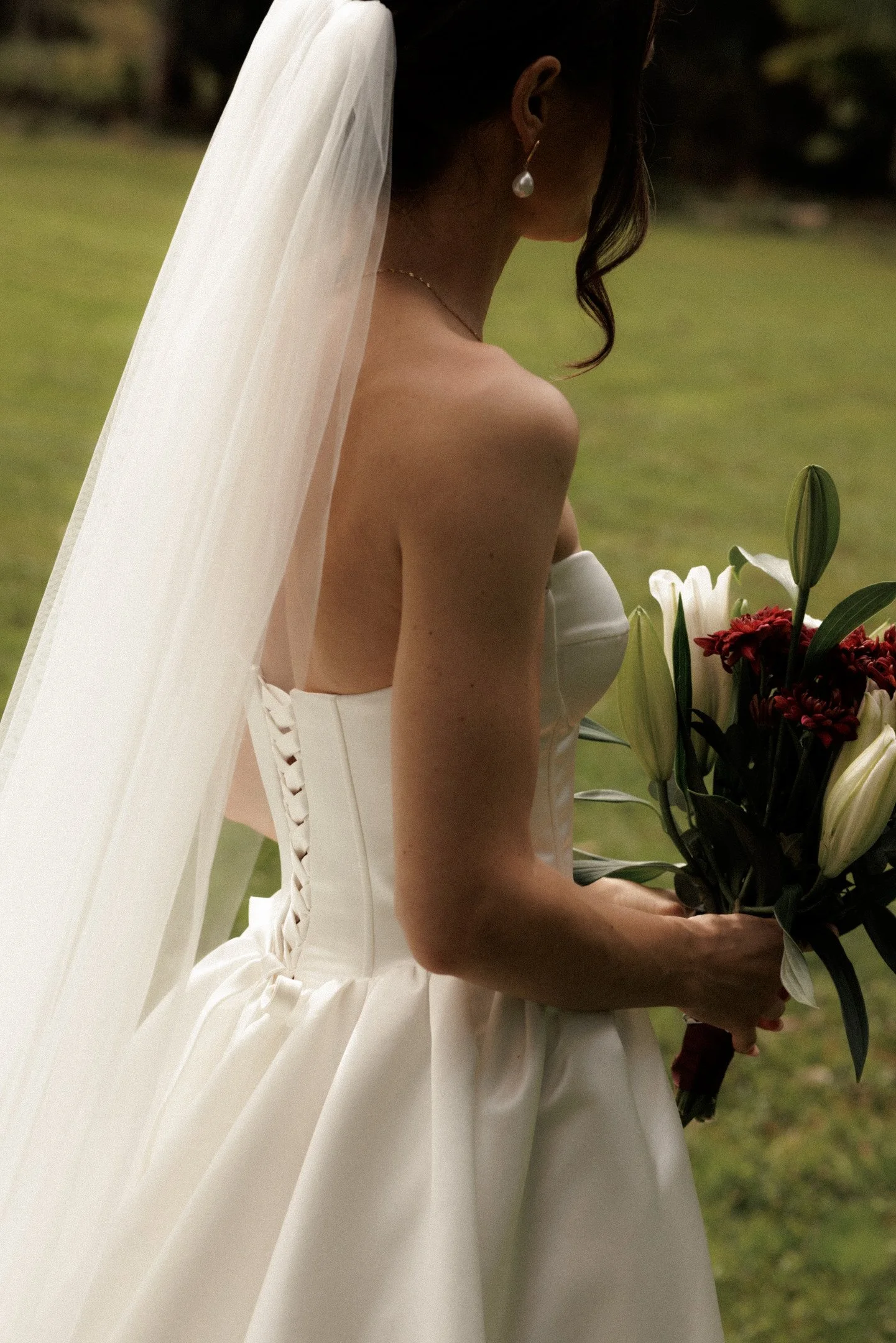 A bride in a white wedding dress with a veil holding a bouquet of white and red flowers in an outdoor setting.