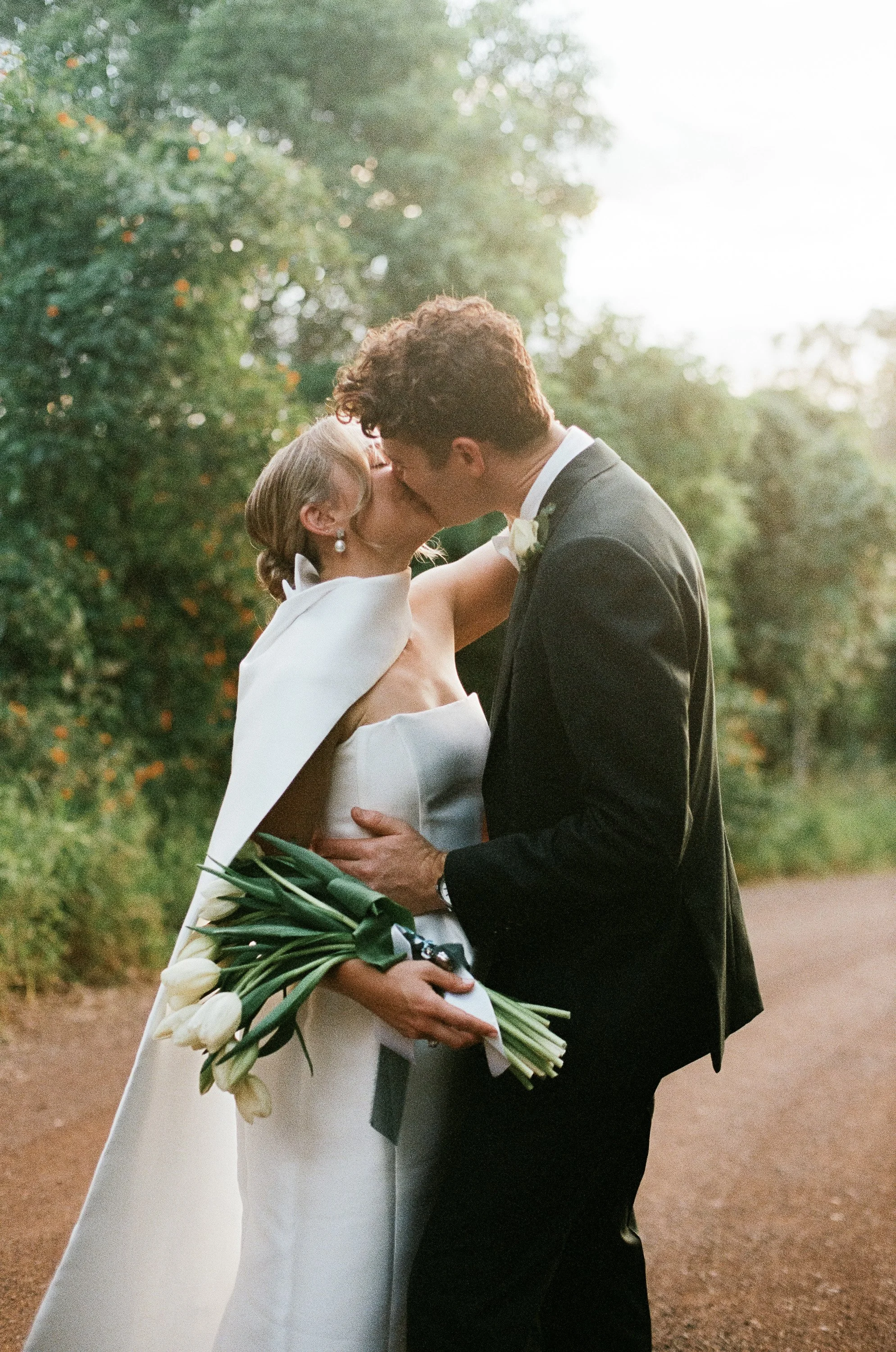 A bride and groom kiss outdoors, with the bride holding a bouquet of white tulips, surrounded by green trees and soft sunlight.