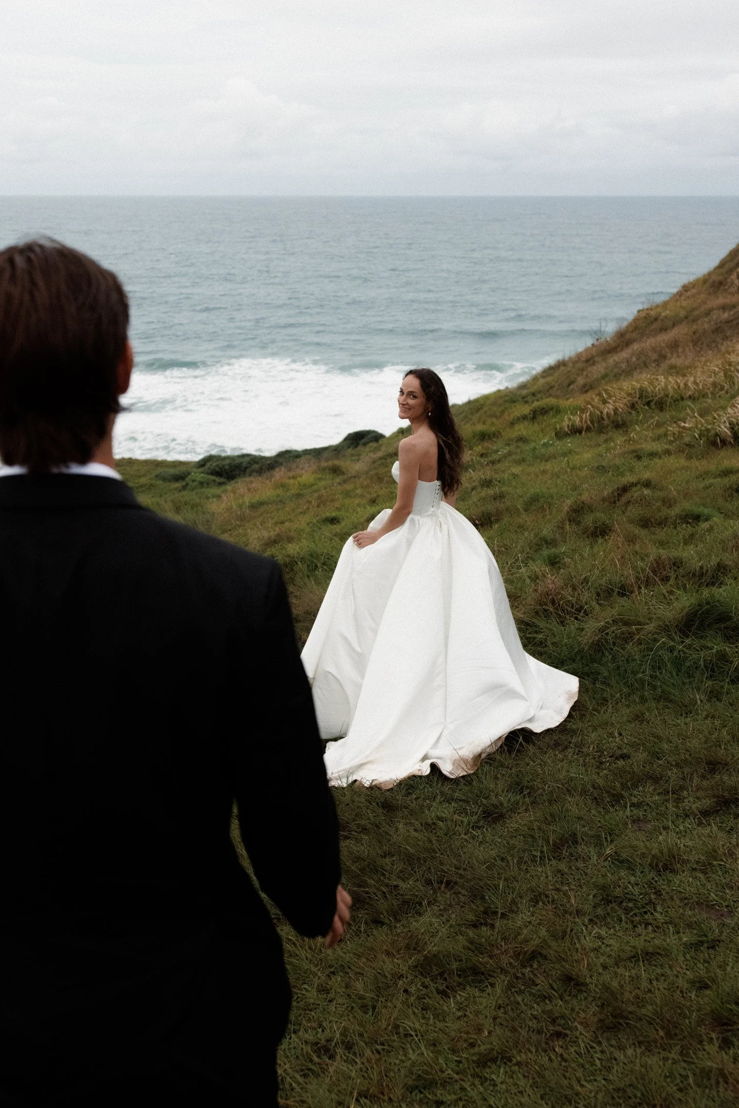 A woman in a white wedding dress sitting on grassy hillside by the ocean with a man in a black suit standing nearby, looking at her.