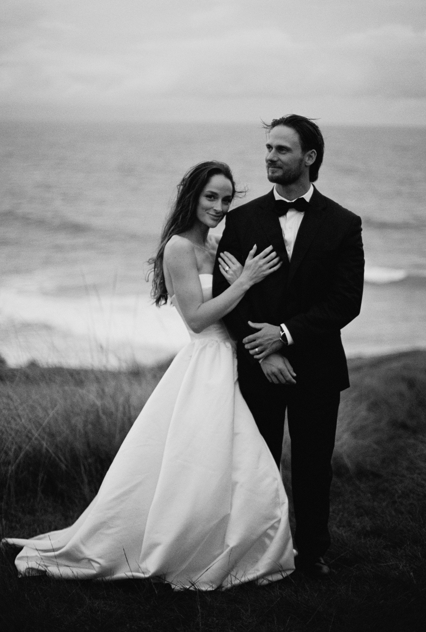 A black and white photo of a bride and groom standing together outdoors, with the bride smiling and the groom looking to the side, near a body of water and grassy field.