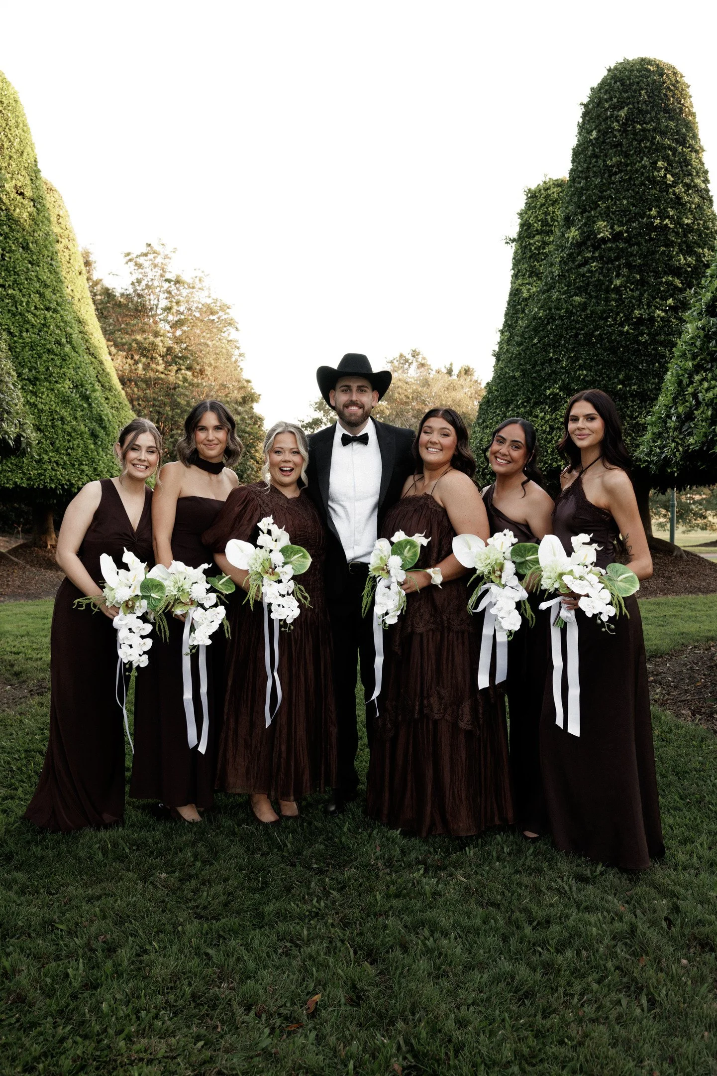 A group of six women and one man standing outdoors in a park with green grass and tall trees, dressed in formal dark brown dresses and a black suit with a bow tie and cowboy hat, holding bouquets of white flowers and smiling.