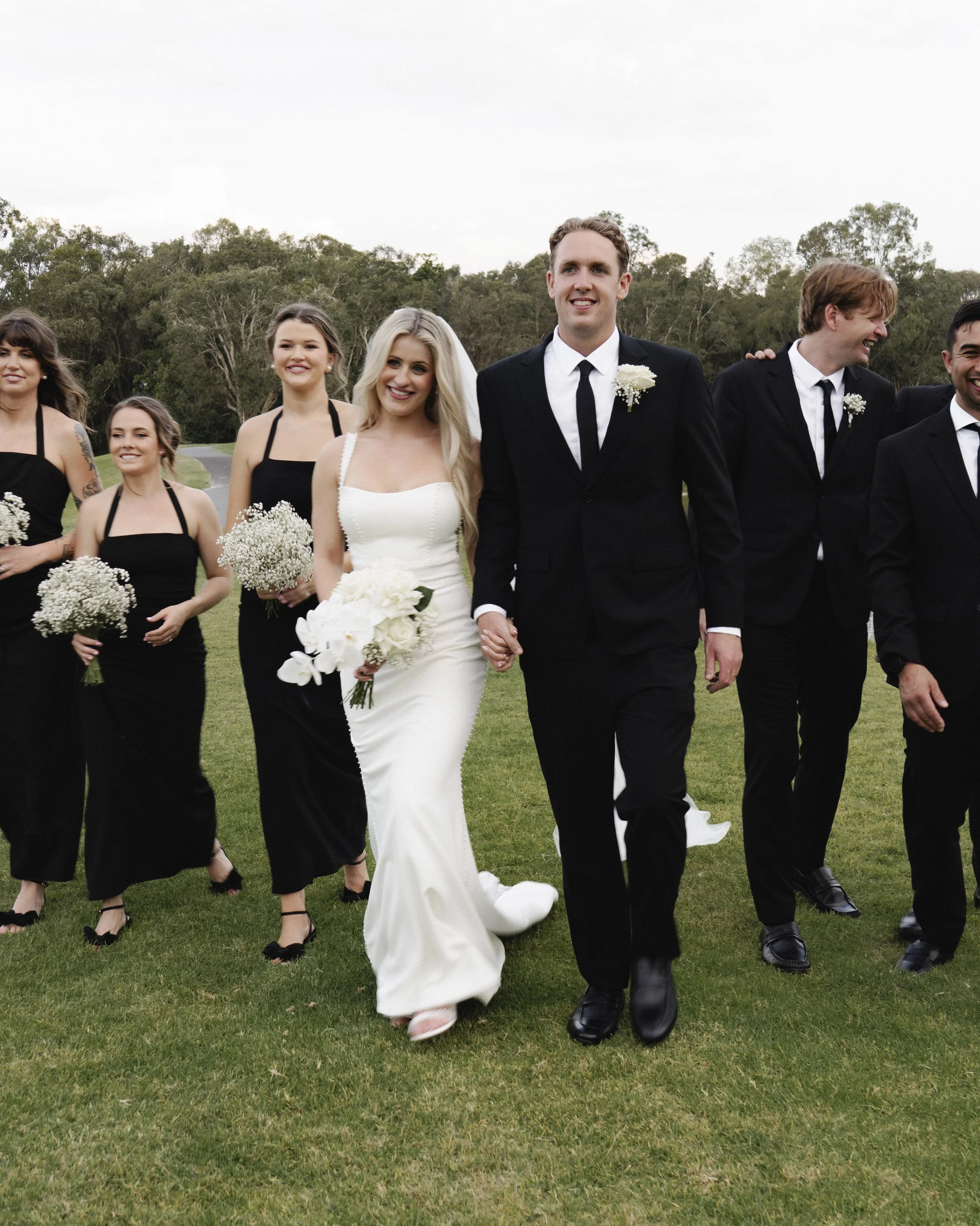A wedding party walking on grass, featuring a bride in a white gown and groom in a black suit, accompanied by bridesmaids in black dresses and groomsmen in black suits, all holding bouquets of flowers - Elevated Weddings shot on 35mm film