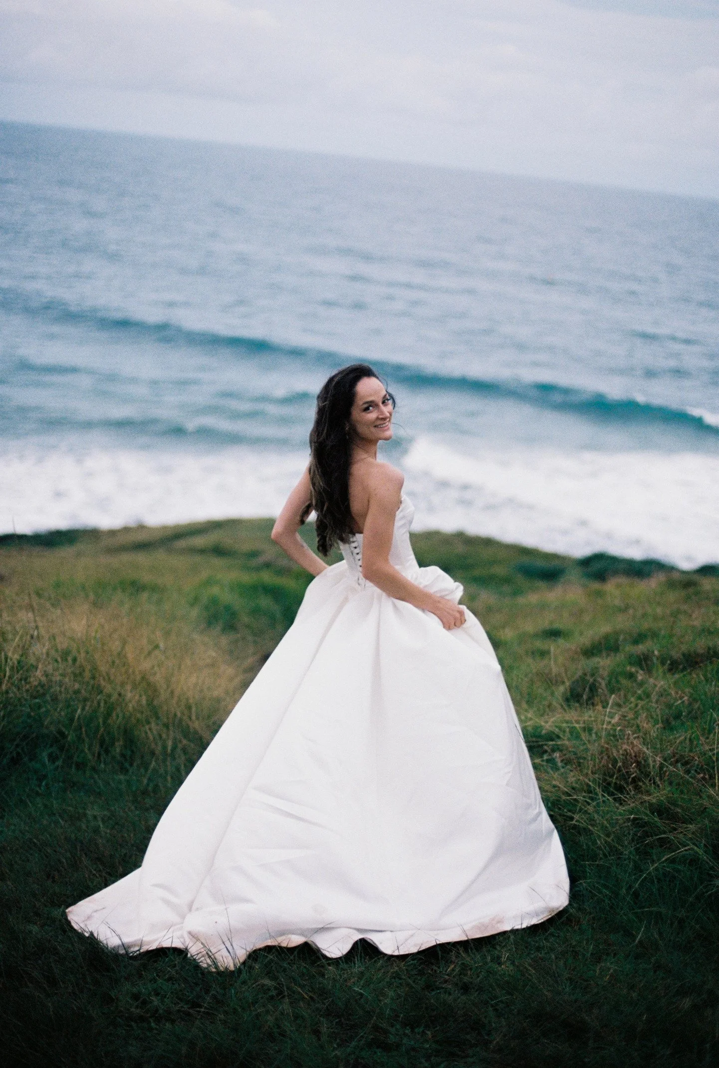 A woman in a white wedding dress smiling outdoors near the ocean with waves and a cloudy sky in the background.