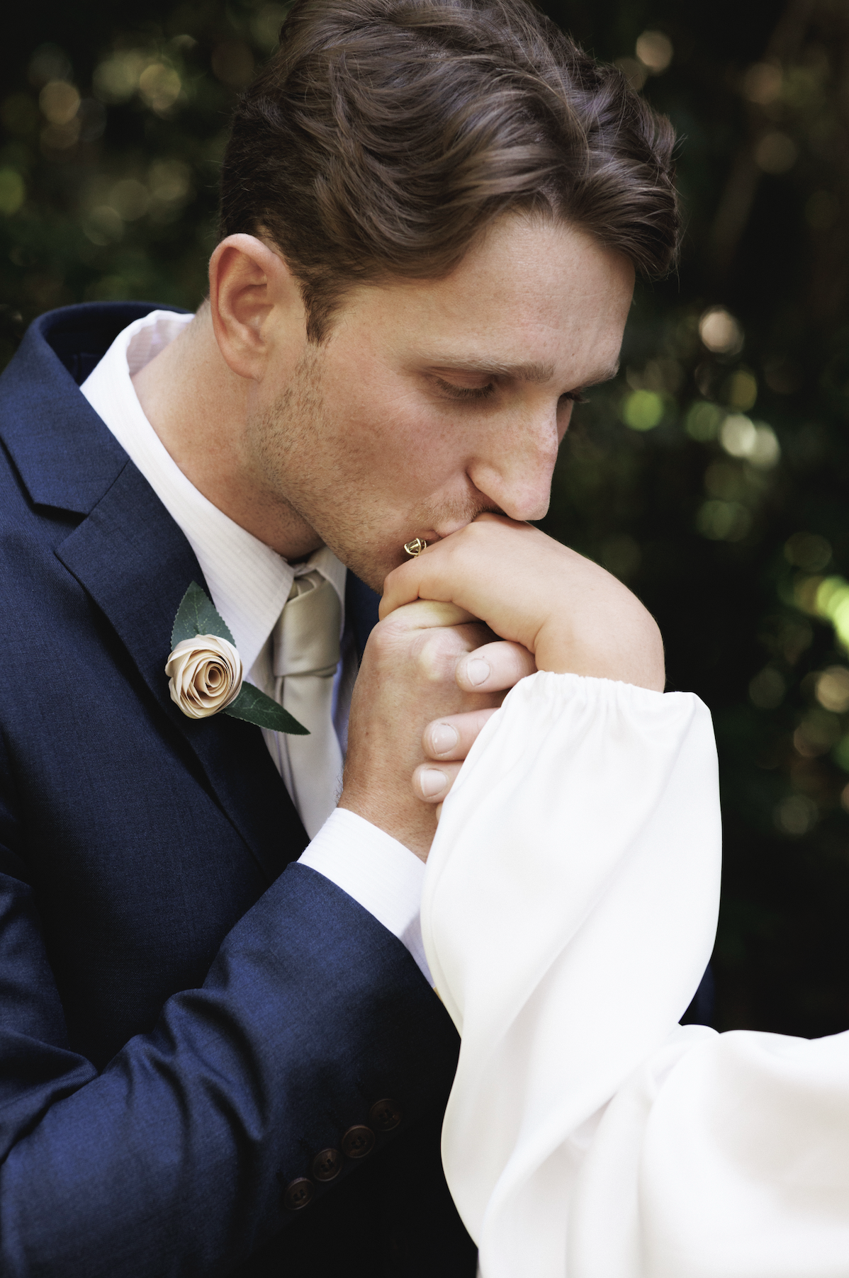 A man in a navy suit kisses the hand of a woman dressed in white, with an outdoor blurred background.