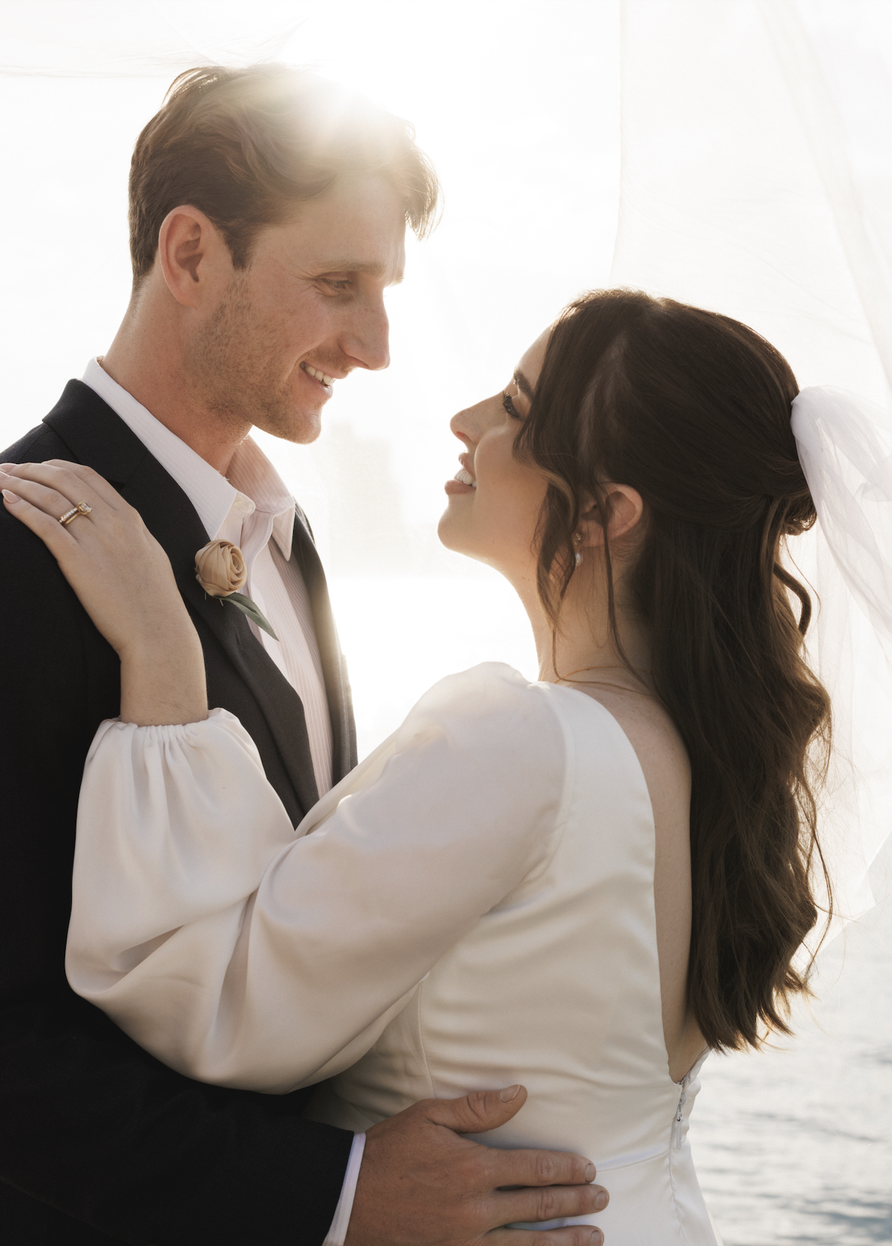 A bride and groom looking at each other lovingly, with the bride's arms around the groom's shoulders and the groom's hand on the bride's waist, standing in front of a bright window.