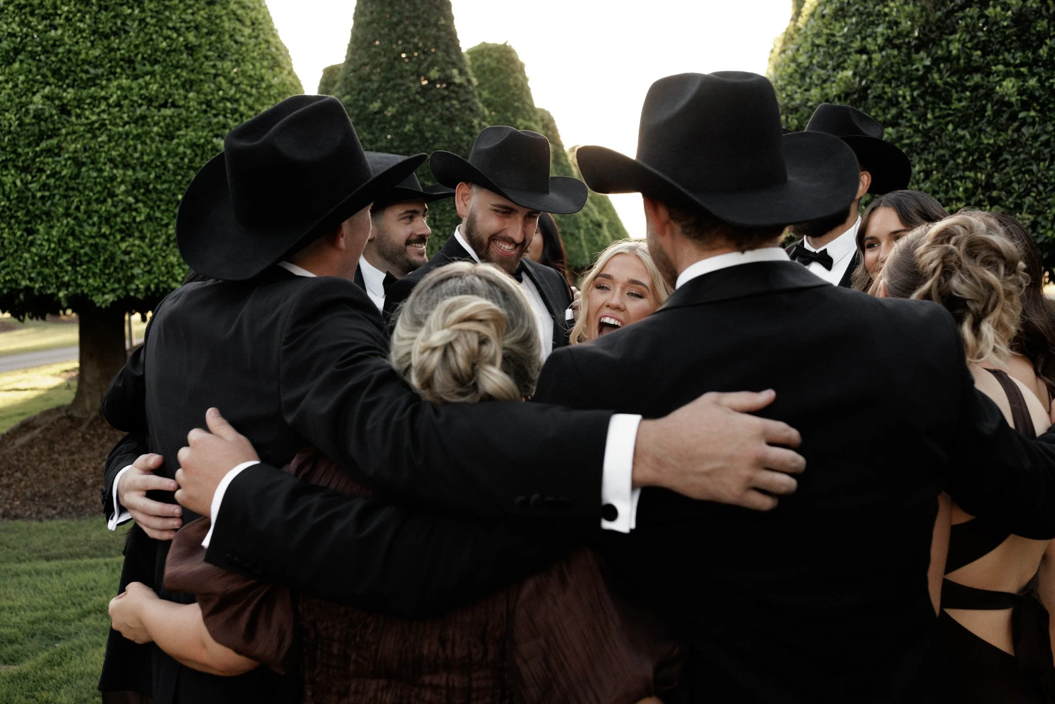 A group of people dressed in black tuxedos and cowboy hats gathered together, embracing in a circle outdoors with manicured bushes and trees in the background.