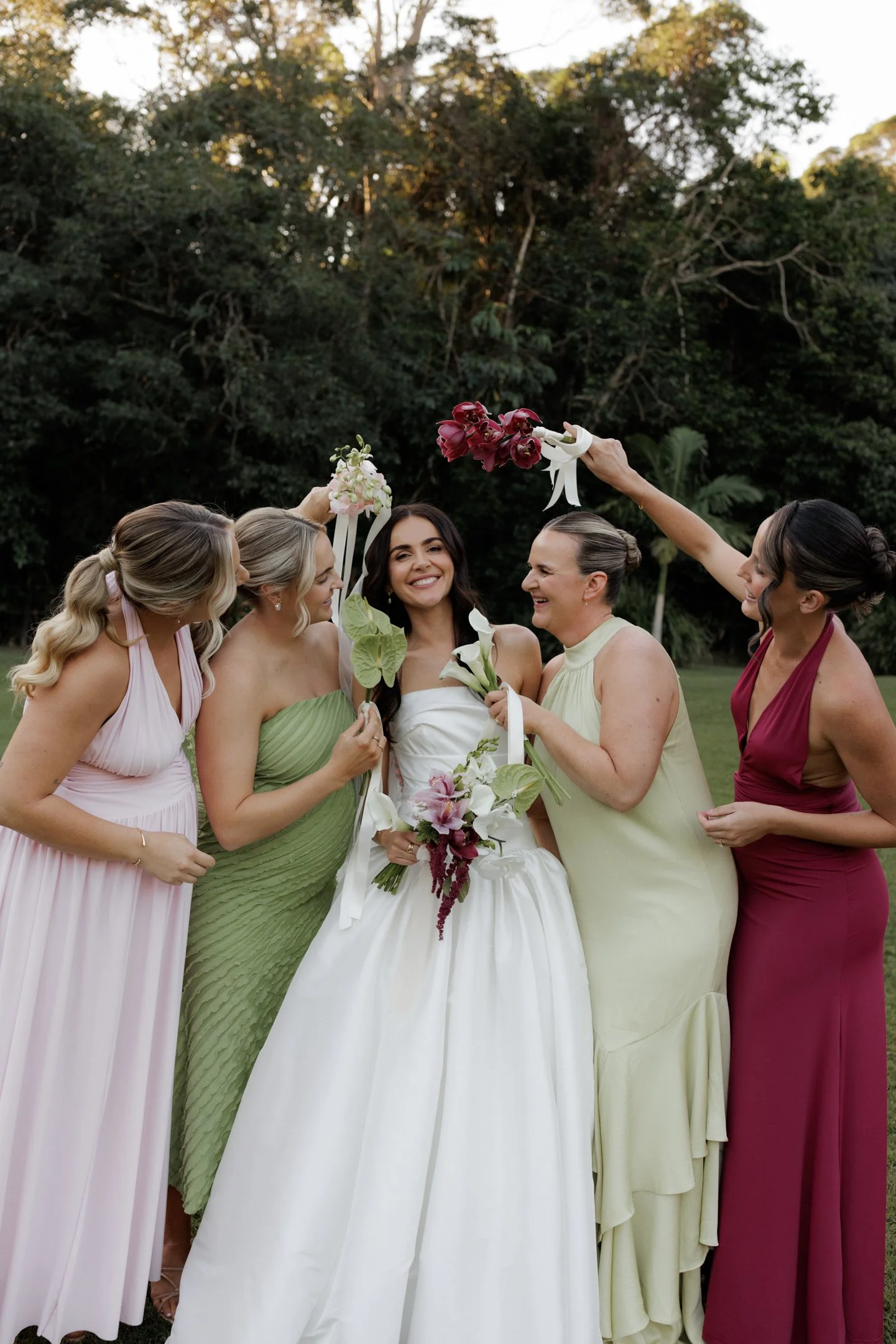 Group of six women in colorful dresses celebrating outdoors with a bride holding a bouquet, while others hold flowers and raise hands, surrounded by greenery.