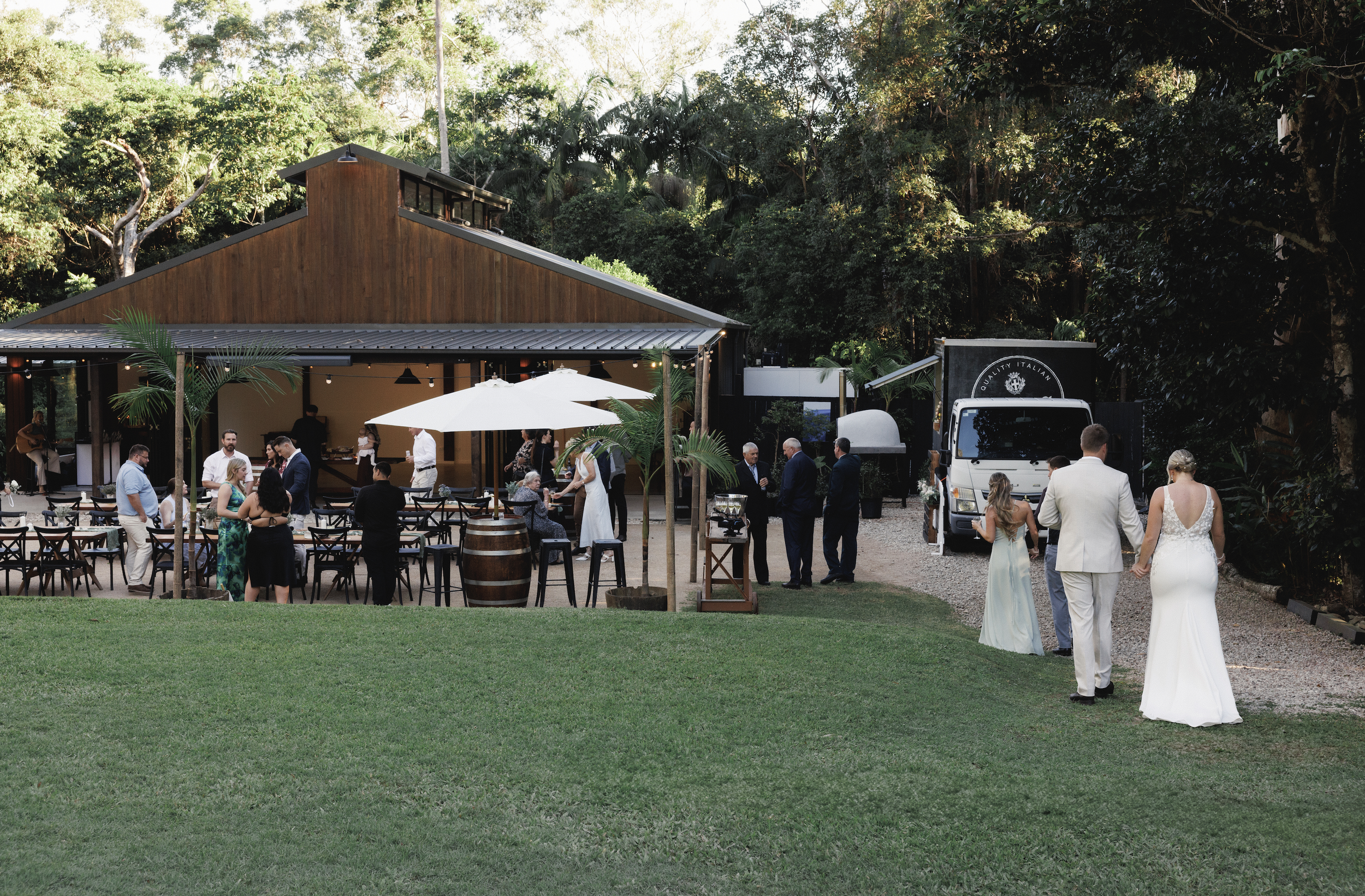 Wedding reception outdoors with guests in formal attire, a bride in a white wedding dress, and a groom in a white suit walking on a grassy area toward a wooden building, with trees and a food truck in the background.