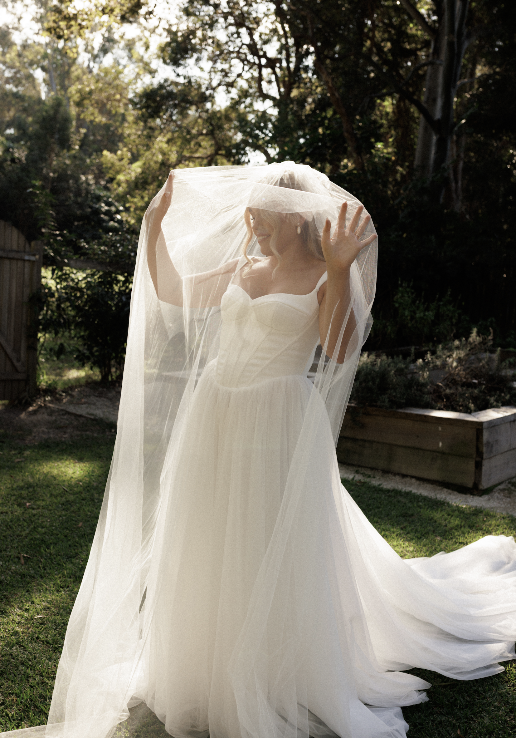 A woman in a wedding dress with a veil standing outdoors, smiling and waving, surrounded by trees and greenery.