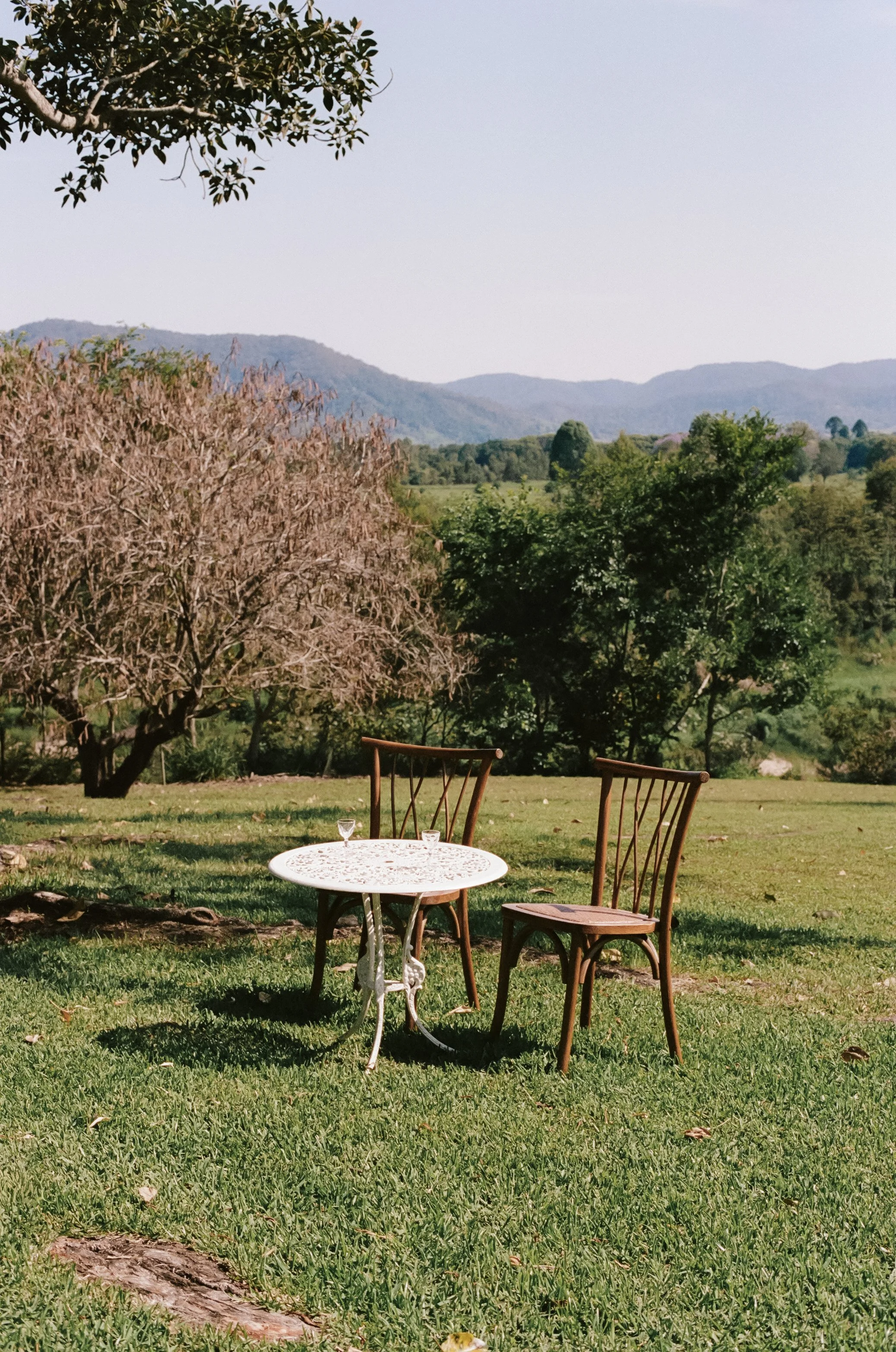 A white metal table with ornate design on a grassy lawn, with two empty wine glasses. Two wooden chairs with vertical slats are positioned around the table. The scene overlooks a lush landscape of trees and rolling hills or mountains in the distance,