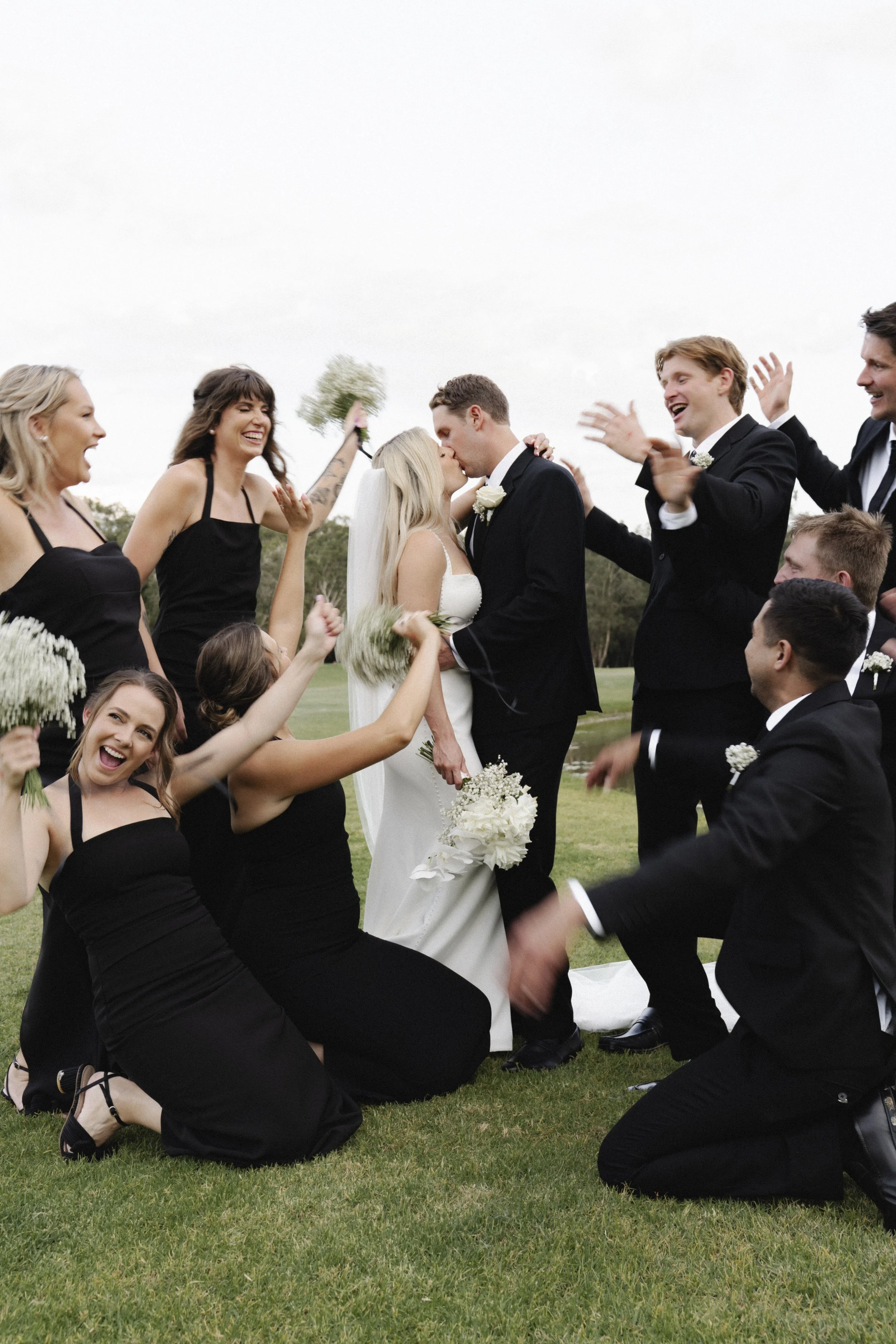A wedding party outdoors on a grassy area, with a bride and groom kissing, surrounded by bridesmaids and groomsmen celebrating and cheering.