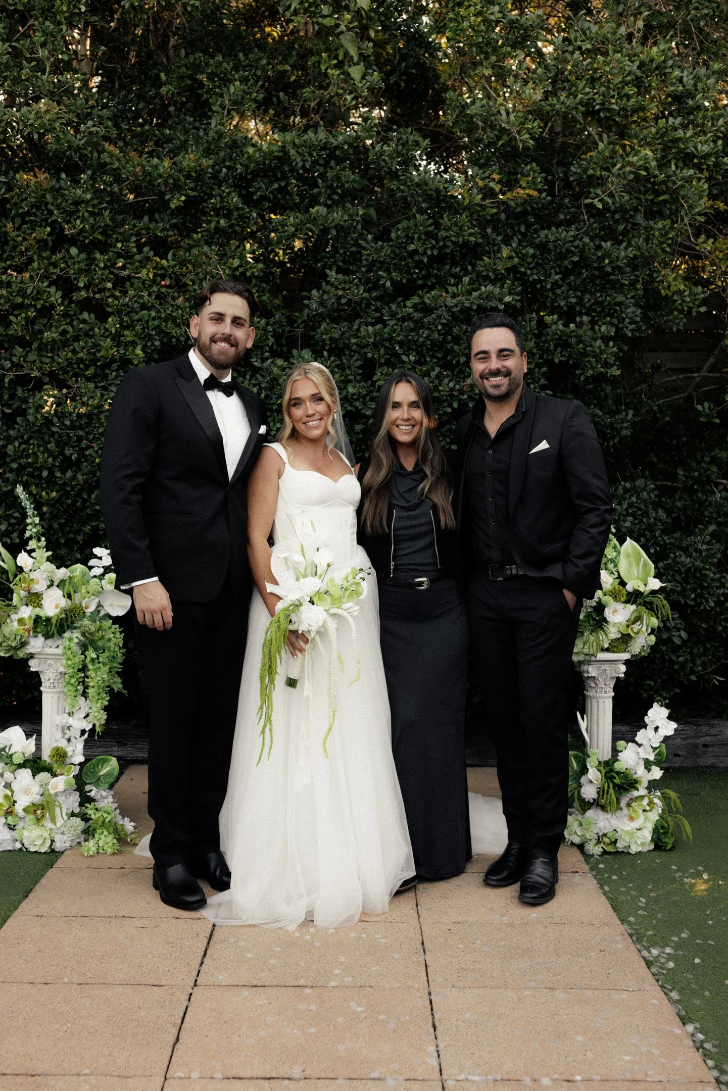 Group of four people standing outdoors in front of a lush green hedge, dressed in formal attire for a celebration or wedding, with floral arrangements and a decorative arch behind them.