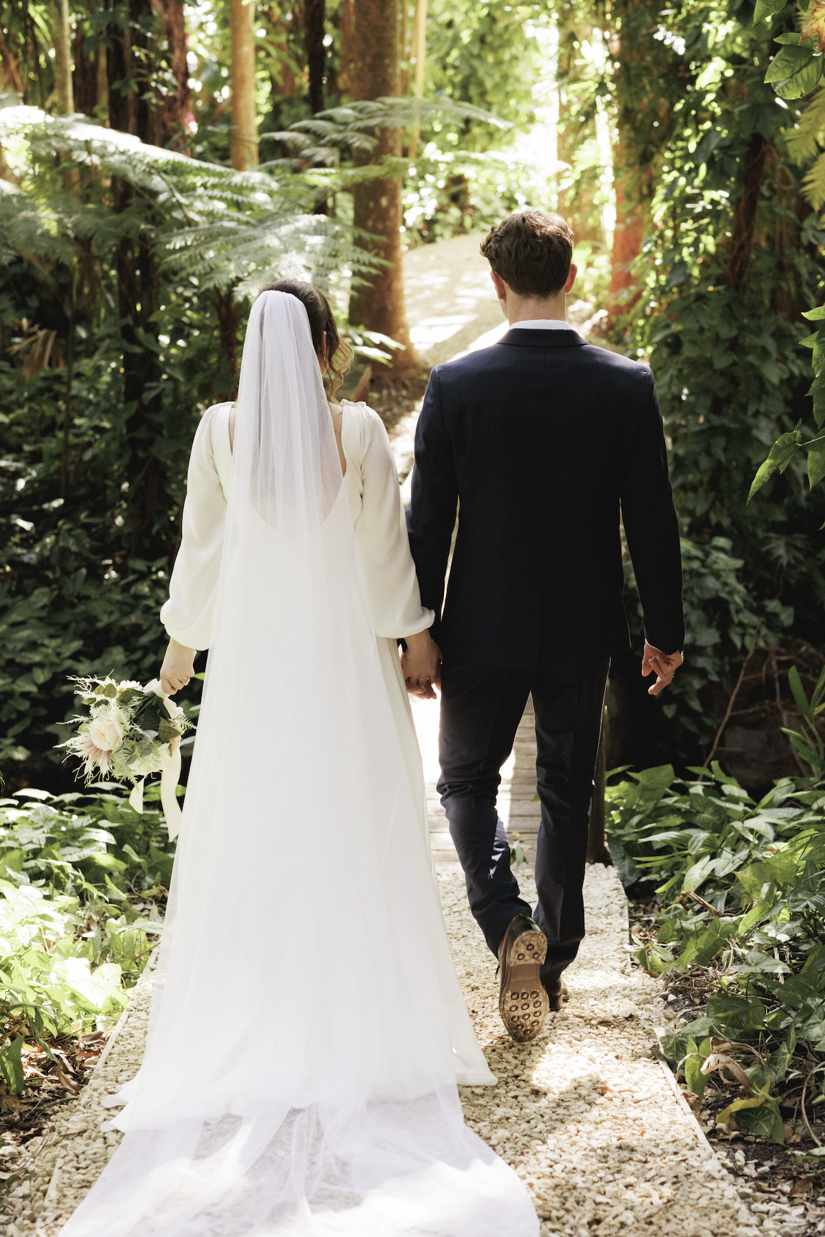 A bride and groom walking hand in hand outdoors in a lush, green forest, with sunlight filtering through trees.