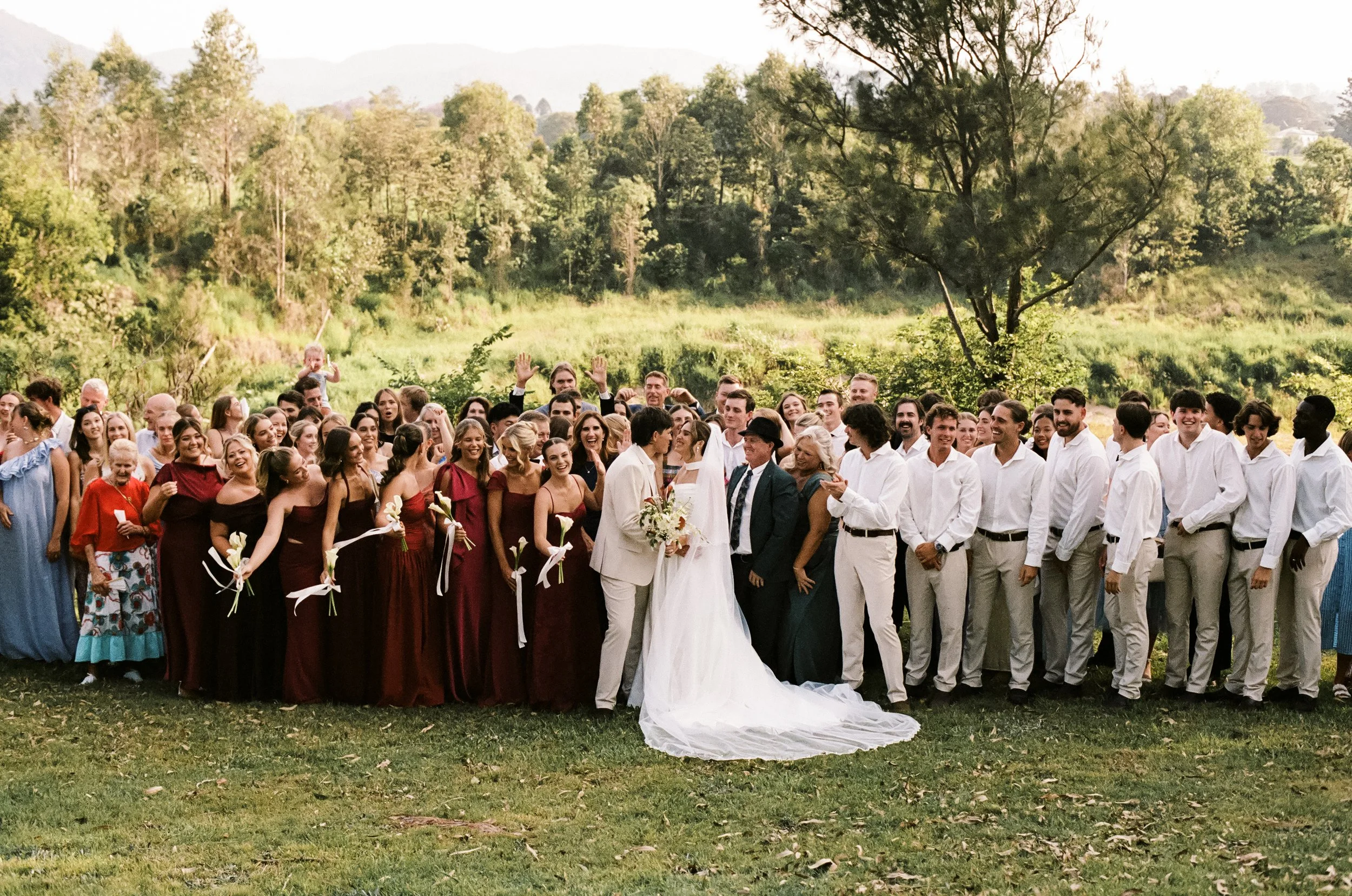 A large wedding party outdoors with many people, the bride and groom in the center, surrounded by bridesmaids in burgundy dresses and groomsmen in white shirts and gray pants, celebrating in a grassy field with trees and hills in the background.