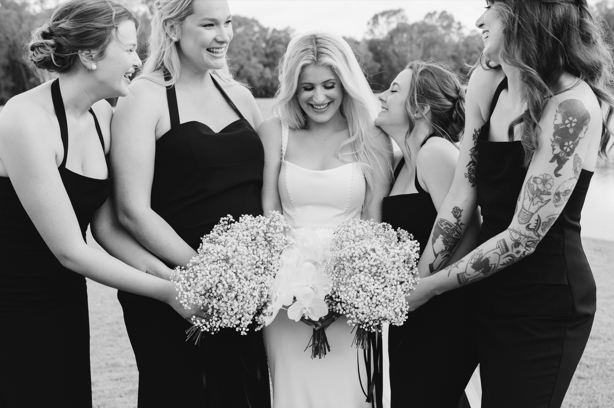 A bride with long blonde hair smiling with four bridesmaids, two with tattoos, all holding floral bouquets outdoors.