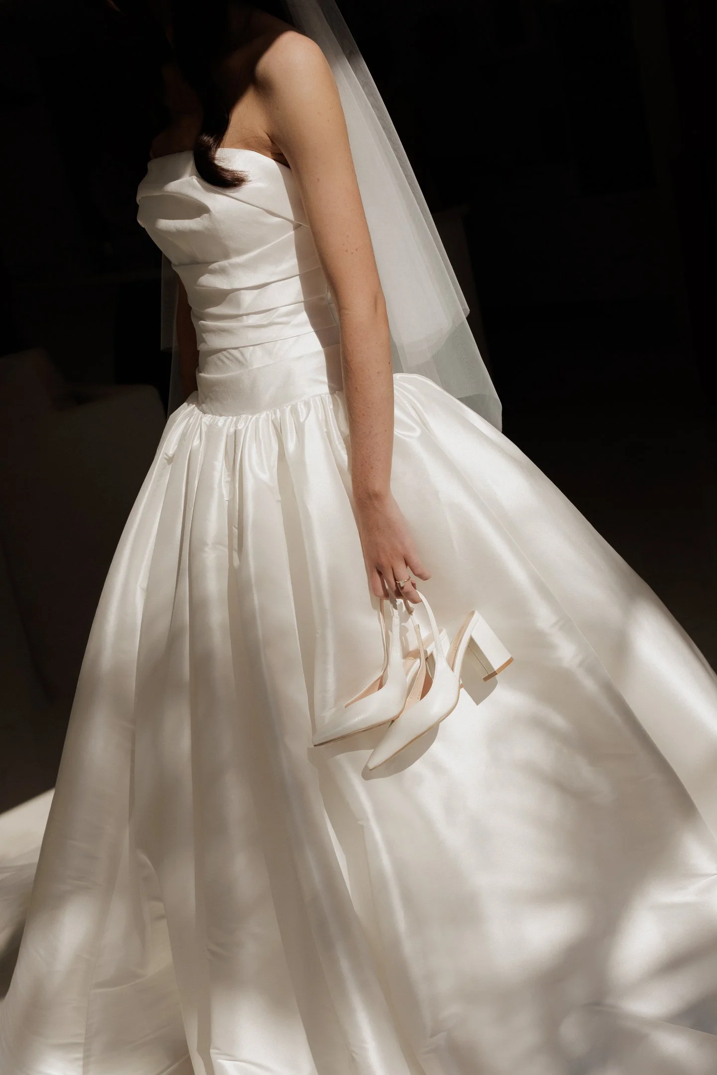 Close-up of a bride in a white wedding dress holding white high-heeled shoes in her right hand and wearing a wedding veil.