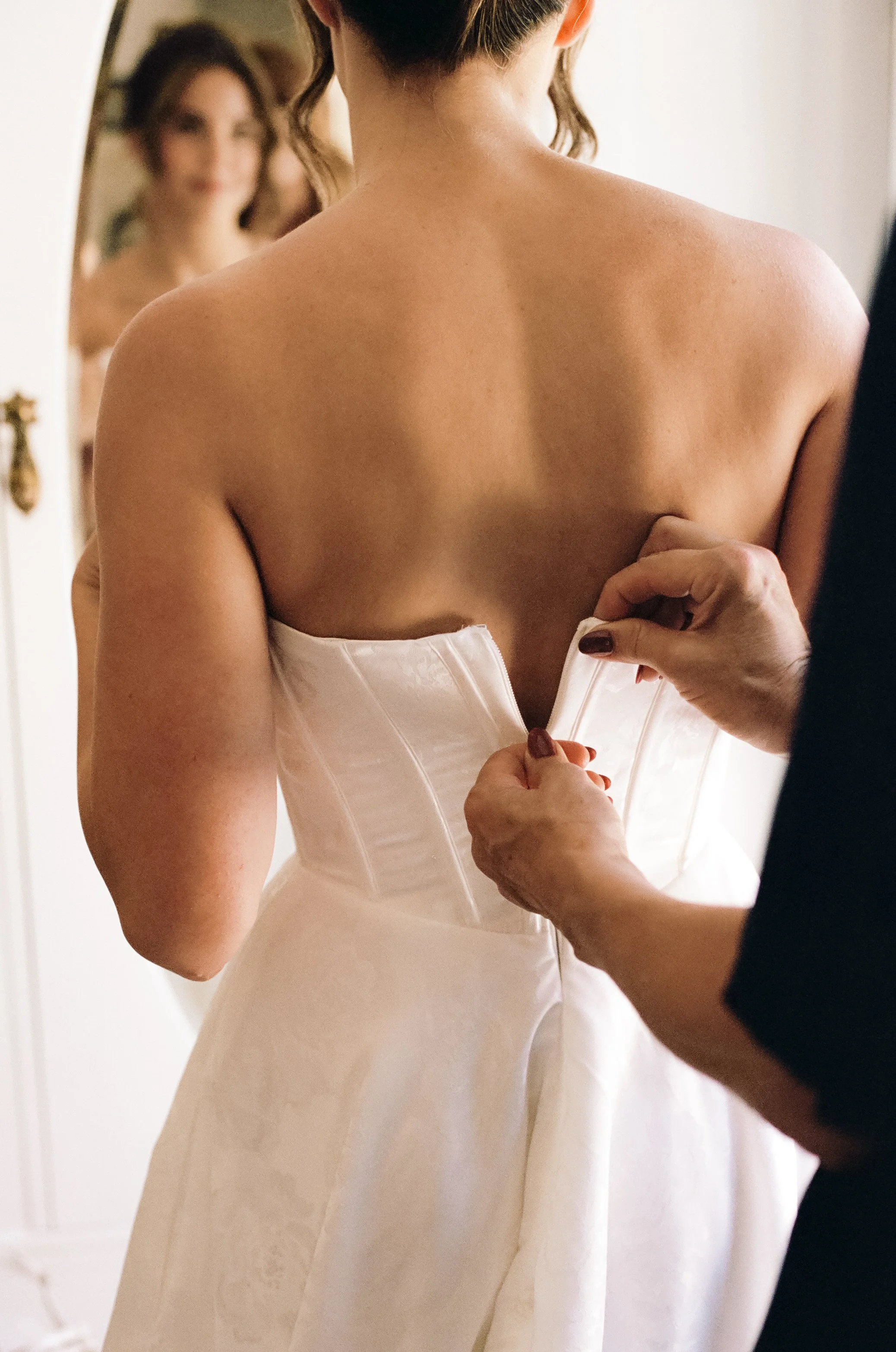 A woman in a white satin wedding dress is being assisted with the back zipper by another person.