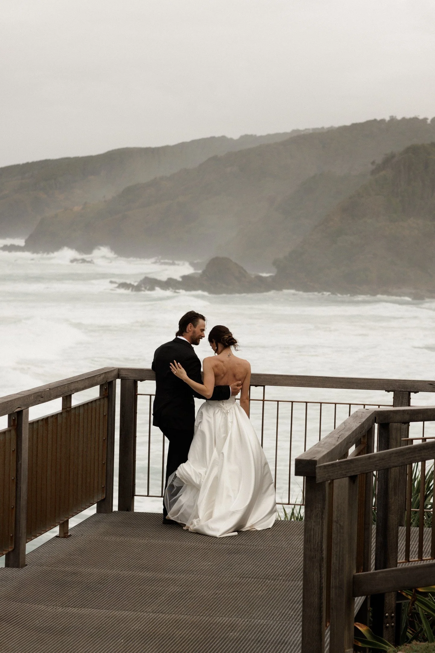 A wedding couple standing on a wooden deck overlooking the ocean on a foggy day, with green hills in the background. The bride is wearing a white wedding gown, and the groom is in a black suit. They are facing each other, holding hands, and smiling.