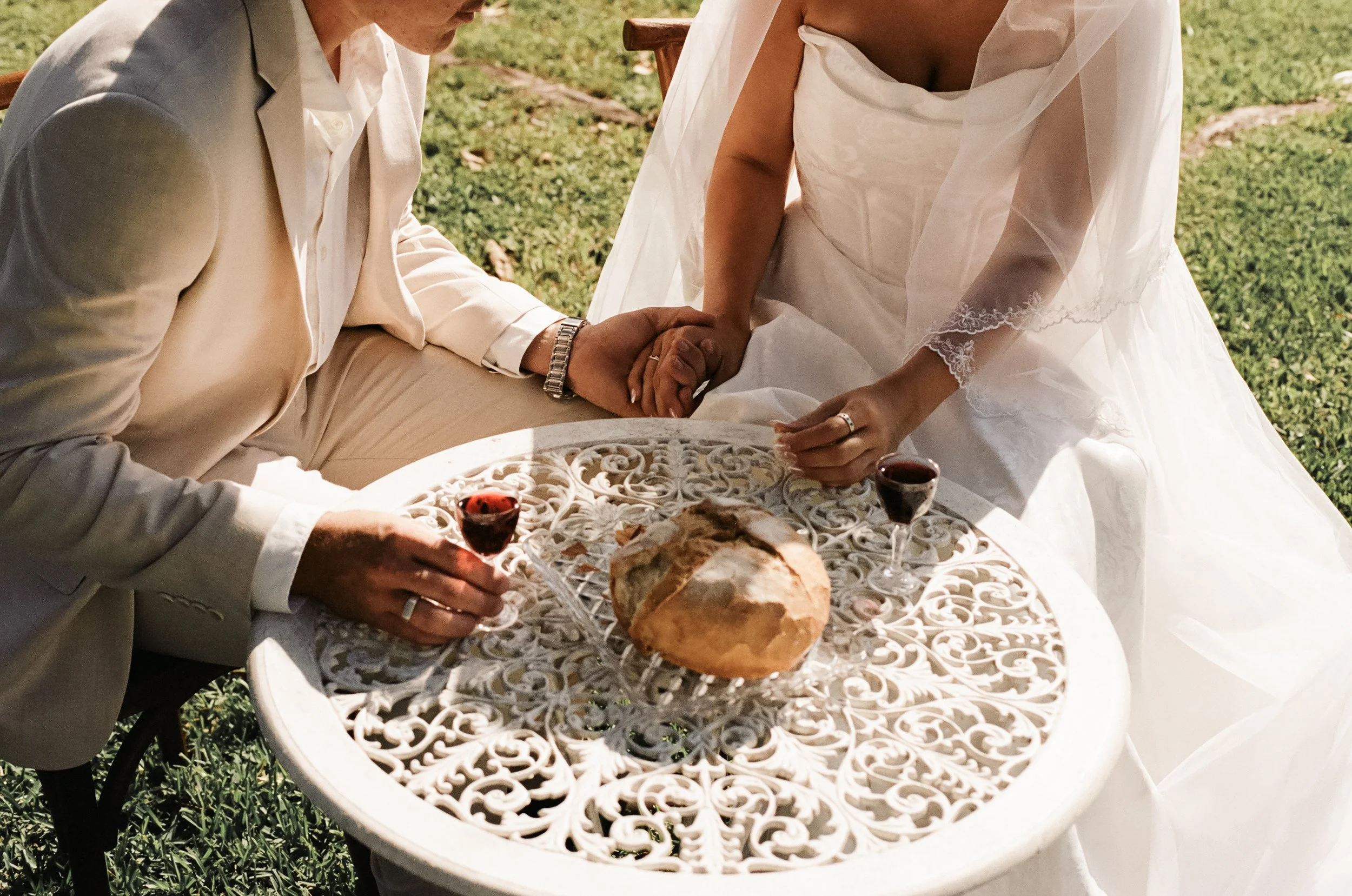 Bride and groom sitting at a decorative white metal table with a round loaf of bread and two glasses of red wine, holding hands in an outdoor setting.