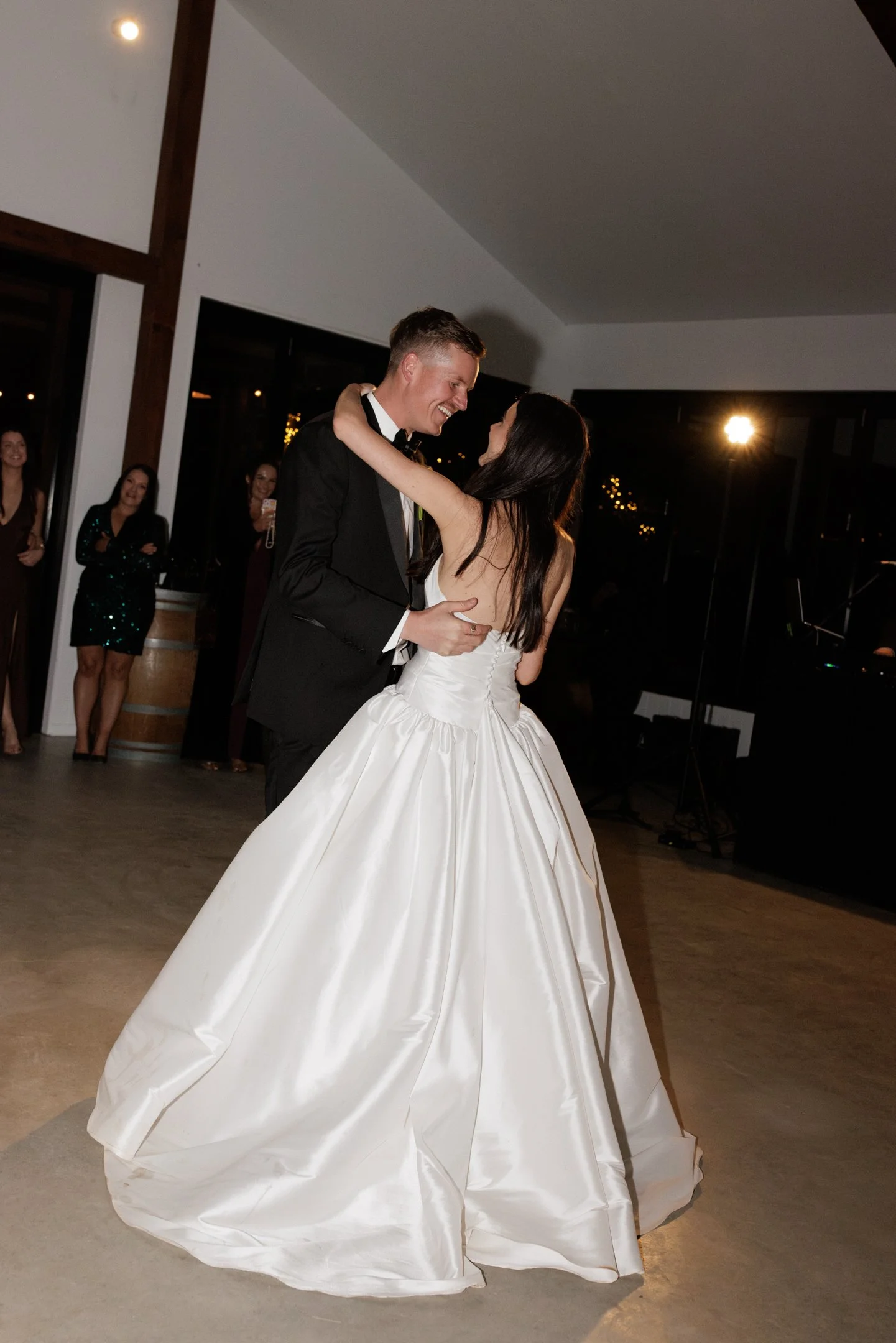 A bride and groom dancing at their wedding reception, with the bride wearing a white, full-skirt wedding gown and the groom in a black tuxedo. Several guests, mostly women, are visible in the background, watching and smiling.